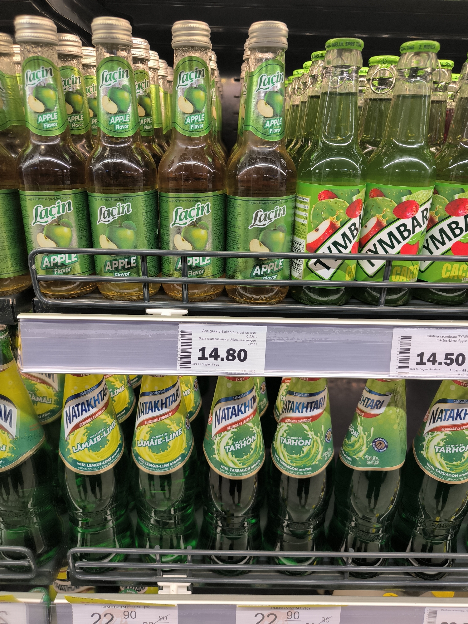 A shelf in a grocery store displays rows of green glass bottles of sparkling drinks. In the foreground, there are several bottles of "NATAKHTARI Georgian Lemonade" in flavors like "LÂMÂIE-LIME" and "TARHON". Behind these, slightly out of focus, are bottles of "LACIN Apple Flavored Sparkling Drink" and "TYMBAR Cactus-Lime-Apple". A price tag on the shelf indicates "14.80" for the "APAC gazate sucul cu gust de Mar" and "14.50" for the "Bautura racoritoare TYMBAR Cactus-Lime-Apple". The environment appears to be a well-lit supermarket aisle.