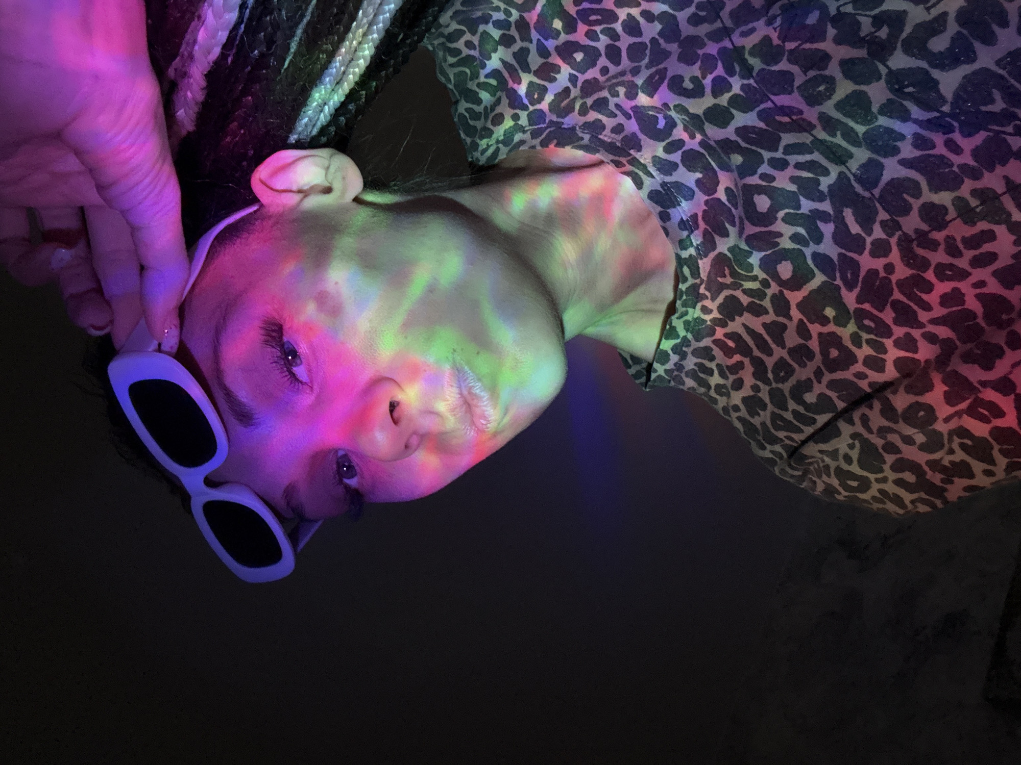 A person lies on their back. The person is wearing sunglasses and a leopard-print top. A hand is reaching toward the person's head. Their face is illuminated with a rainbow of colors. The background is dark. The location is unknown and cannot be determined from the image.
