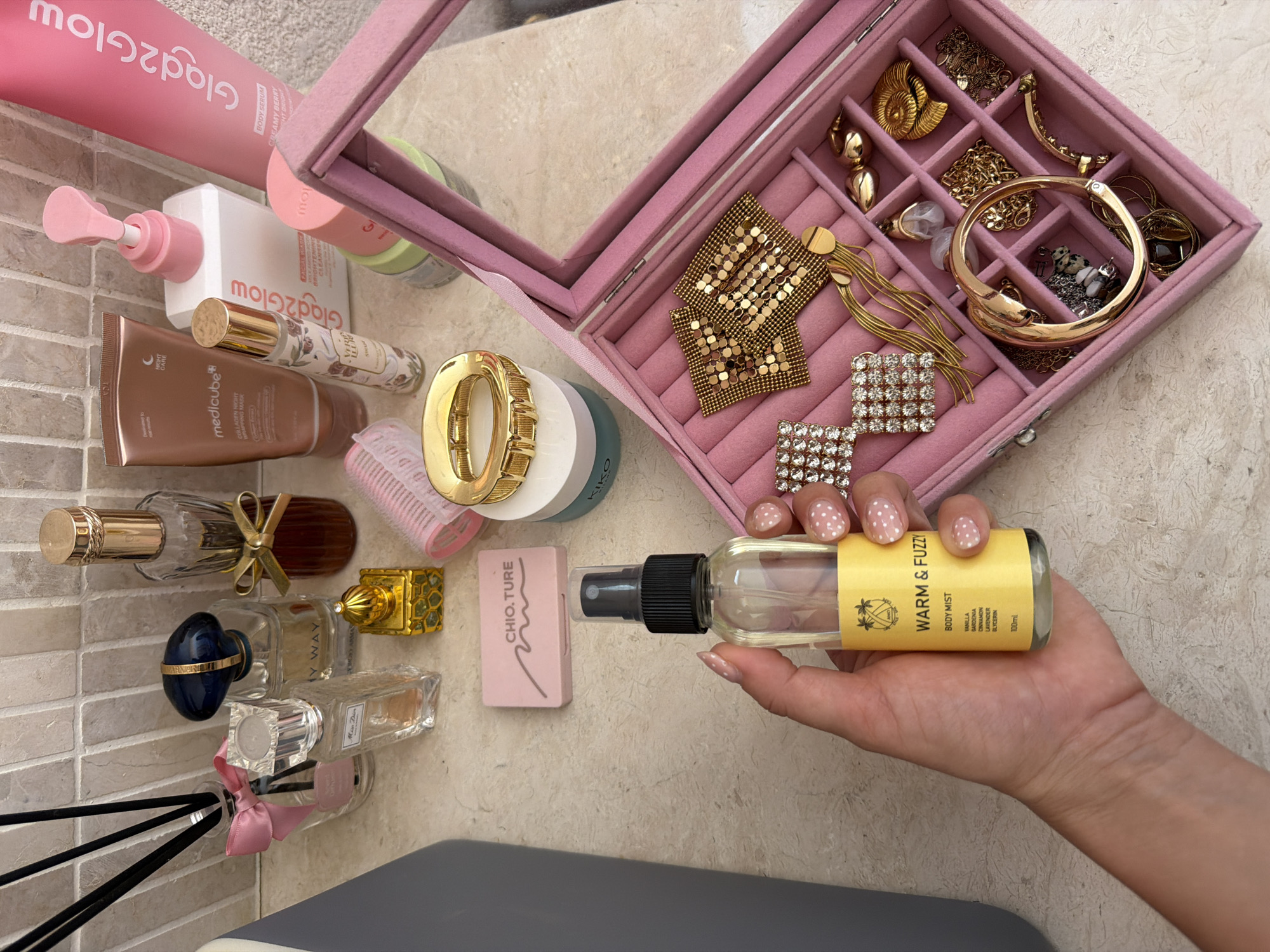 The image shows a flat lay of various beauty products and jewelry on a beige, textured surface, likely a bathroom counter. In the foreground, a person's hand with manicured nails is holding a clear spray bottle labeled "WARM & FUZZY BODY MIST." To the right of the spray bottle is an open pink jewelry box filled with gold-toned necklaces, earrings, bracelets, and rings.
Scattered behind the jewelry box are other beauty items. These include several perfume bottles, a pink hair roller, a tube of "medicube COLLAGEN NIGHT WRINKLE MASK," a pink pump bottle, and two taller pink bottles labeled "Glad2Glow." Further back, a glass diffuser with black reeds and a pink bow is visible. Also present are small, decorative items like a golden miniature building and a pink compact. The background appears to be a tiled wall. The overall impression is a collection of personal care and adornment items.