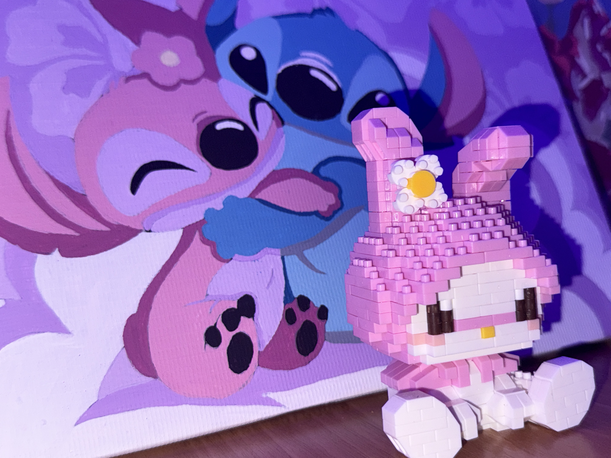 The image depicts a close-up, eye-level shot of a small, pink, pixelated toy figure resembling a character with bunny ears, a flower decoration, and a yellow nose, positioned in the foreground on a wooden surface. Behind the toy, a painting on canvas is visible, featuring two cartoon characters: a pink, feminine character embracing a blue, alien-like character. The painting is rendered in shades of pink, purple, and blue, with the characters appearing to be in a close embrace. The lighting in the scene has a purple hue, likely from ambient light or a specific light source. The background is blurred and consists of the purple-tinted canvas. The image does not contain any recognizable landmarks or elements that would indicate its location in Rayevskiy, Russia.