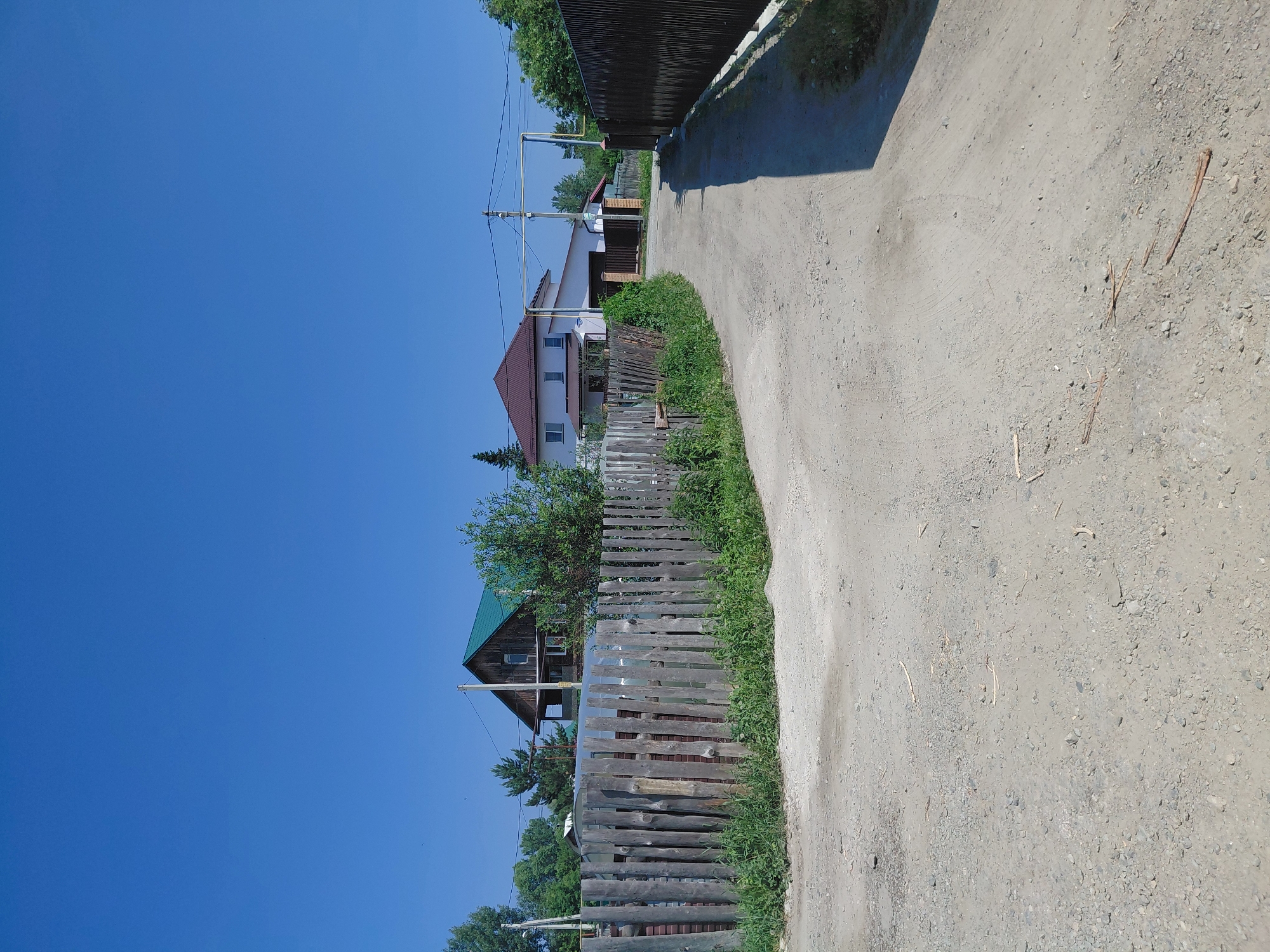 The image depicts a dusty, unpaved road winding through a rural or suburban area. The road surface is a mixture of dirt and small gravel. On the left side of the road, a rustic wooden fence constructed from vertical logs borders the property. Beyond the fence, a two-story wooden house with a green roof and a dark wood exterior is visible, partially obscured by trees. Further down the road on the left, a modern, two-story white house with a brown roof is visible, featuring multiple windows and a garage.
On the right side of the road, a dark, solid fence runs along the property line, casting a long shadow onto the road. Overhead, electrical wires are strung between poles, and a metal pipe, possibly a gas line, spans across the road. The sky is a clear, bright blue, indicating a sunny day. The vegetation consists of green trees and overgrown grass along the roadside and behind the fences. The scene appears to be in a settlement where houses are spaced apart, with unpaved roads and a mix of traditional and modern housing. Given the context provided by the prompt, the location is a street in Novosibirsk, Russia.