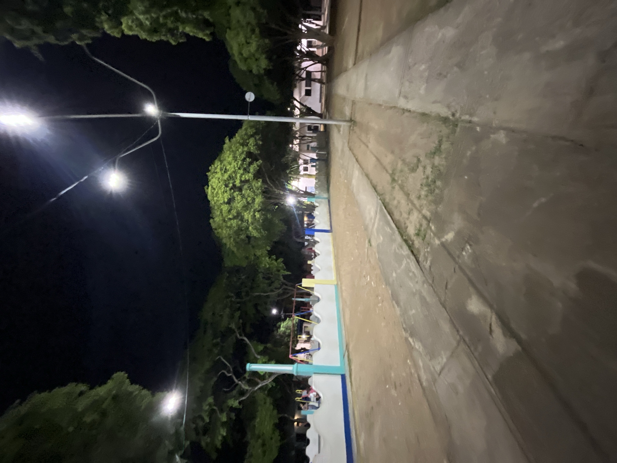 The image depicts an outdoor scene at night, illuminated by streetlights. In the foreground, a paved path runs through a sandy or dirt area. To the left, a low, decorative wall with a wavy top separates the path from a playground area. The playground features swings and what appear to be colorful structures. Trees with dense green foliage are scattered throughout the scene, with some overhead lighting from street lamps visible. In the background, a building with windows is visible, suggesting an urban or semi-urban setting. Based on the available information, this location is identified as a park or recreational area in Santa Ana, Venezuela. The image shows no discernible people or specific actions.