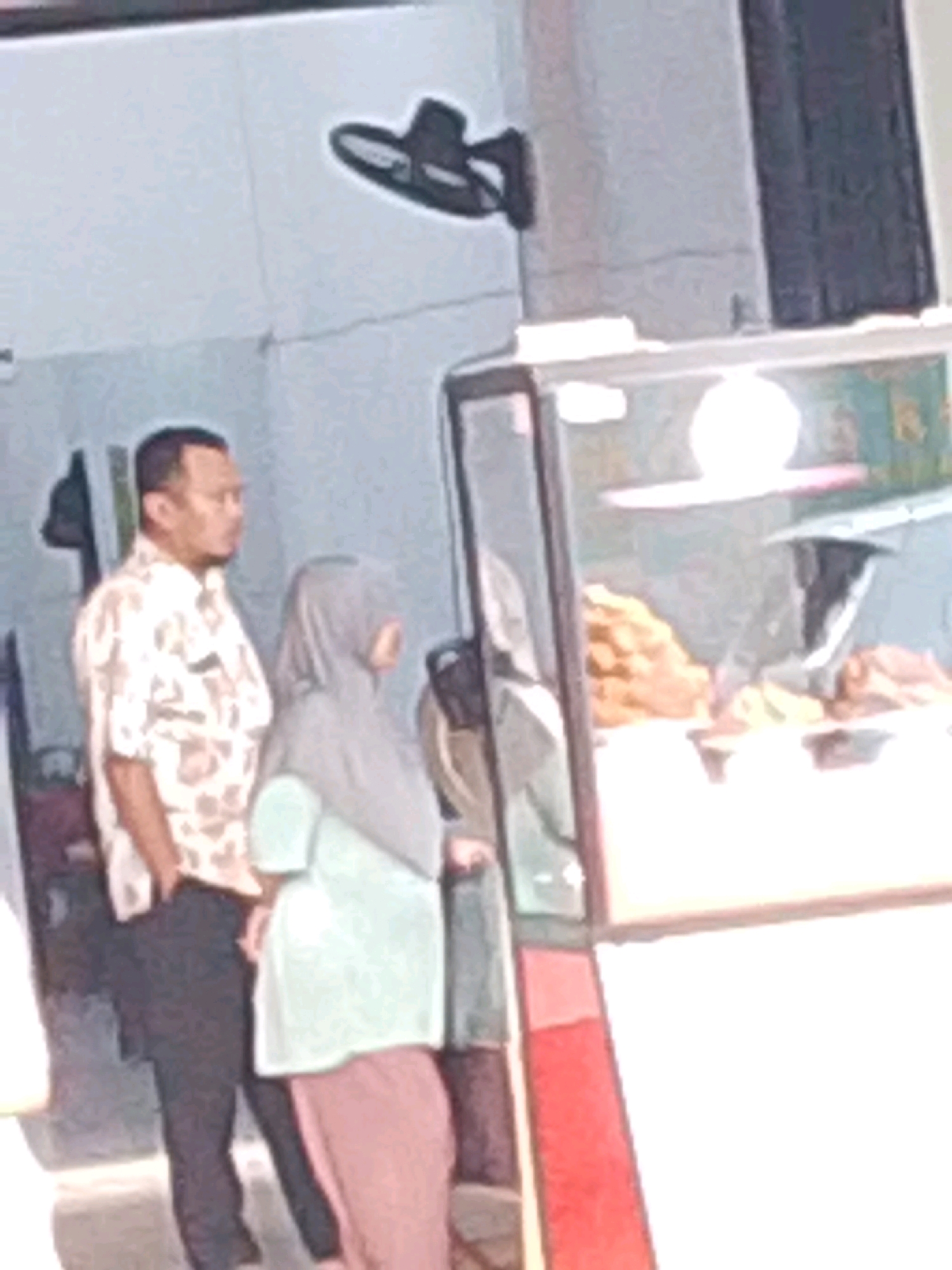 A man in a patterned shirt and dark pants stands to the left of a woman wearing a light green hijab and a pink skirt. The man is facing right, and the woman is facing away from the camera. Behind them, a glass display case contains food items, possibly snacks or pastries, illuminated by a bright light. The background appears to be an outdoor or semi-outdoor setting with a tiled wall and a black fan mounted on the wall above and to the left of the man. The scene is captured in what appears to be a street-level establishment in Probolinggo, Indonesia, likely a food stall or small eatery. The overall image quality is grainy, suggesting it was taken with a low-resolution camera or under suboptimal lighting conditions.