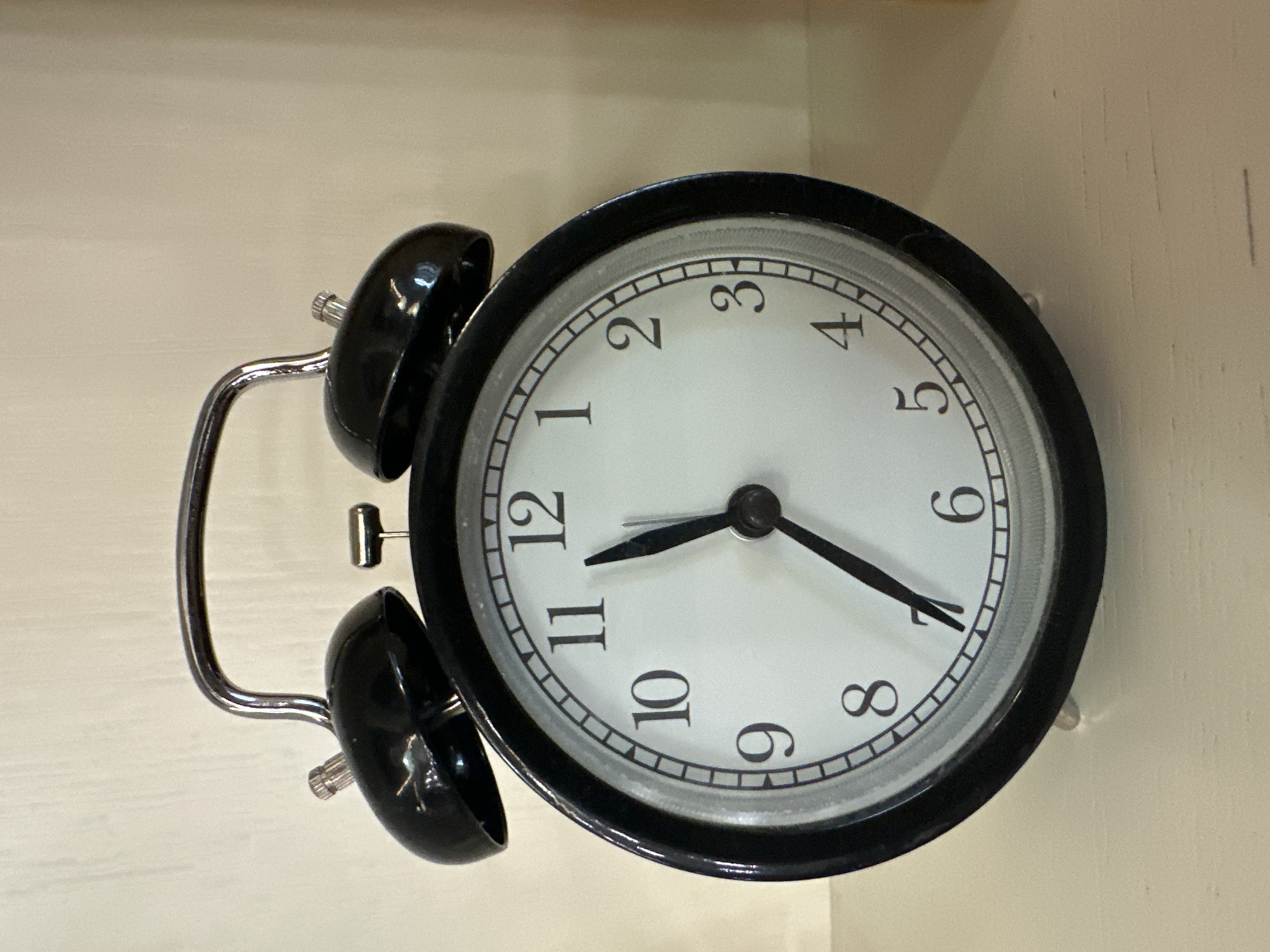 A black alarm clock sits on a light-colored surface. The clock face is white with black numerals. The hour hand points slightly before 12, and the minute hand points at 7. The alarm bells are black and positioned on top of the clock. The scene is indoors, with the clock resting on a flat surface, possibly a shelf or table. The setting appears to be a domestic or office environment. There is no information available from the image that would suggest the location is Moscow, Russia.