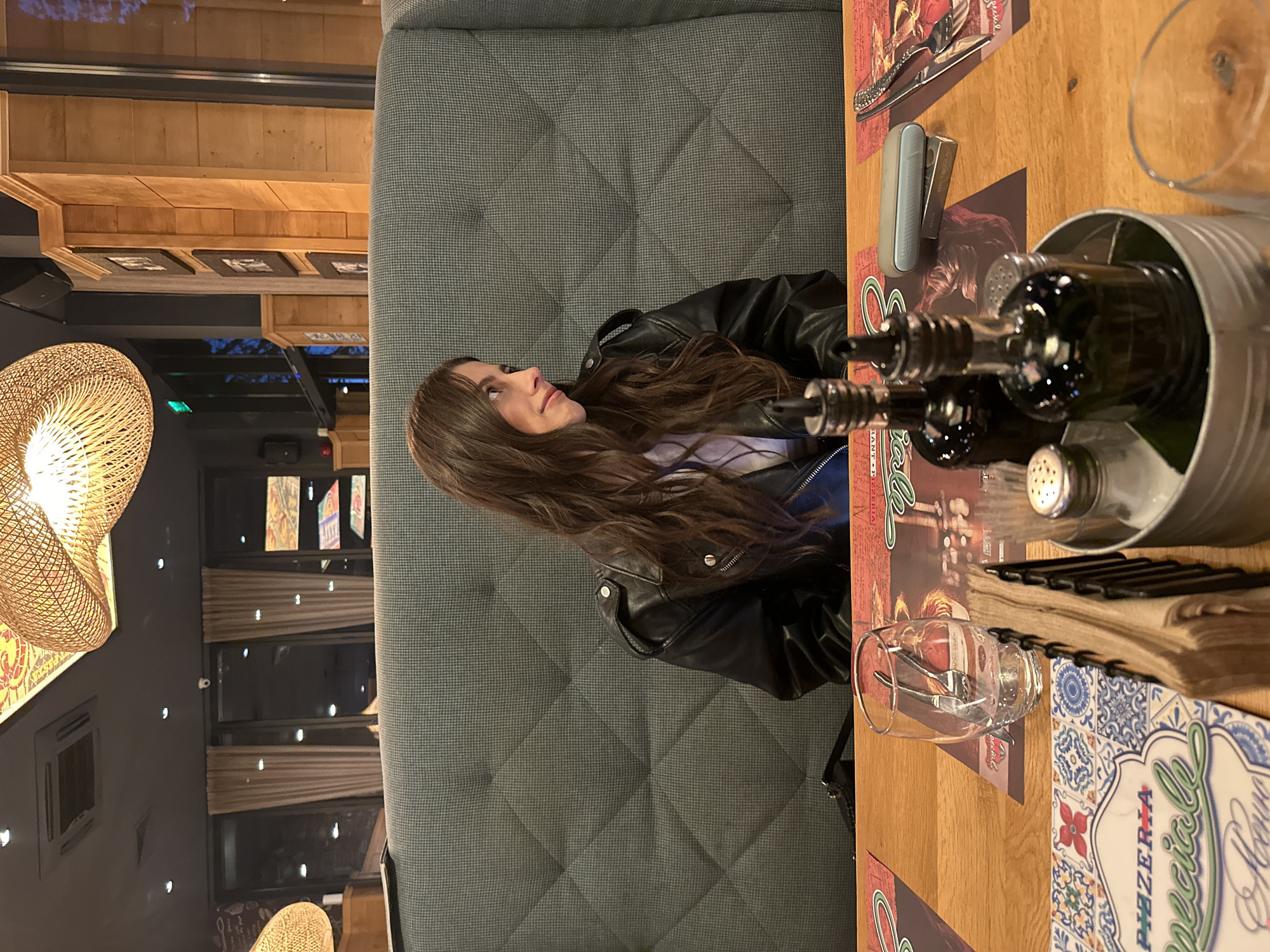 This image depicts an indoor scene within a restaurant or pizzeria, located in Plovdiv, Bulgaria, likely during the evening hours. The primary subject is a woman with long, wavy dark brown hair, wearing a black leather jacket over a light-colored top. She is seated on a grey, quilted upholstered booth, looking upwards and to her left with a serene and slightly smiling expression, her face in profile.
The setting features warm lighting, highlighted by a large, illuminated woven wicker pendant lamp on the left. The interior design incorporates natural wood paneling on the walls, adorned with framed pictures, contrasting with a darker wall section that includes windows, some fitted with blinds, and small recessed lights.
On the wooden table, a casual dining setup is visible. Colorful placemats, one prominently featuring the text "PIZZERIA Special Menu" with decorative blue and red patterns, are laid out. A metal caddy holds three bottles, likely condiments such as oil and vinegar, flanked by salt and pepper shakers. Other table items include an empty clear drinking glass, another glass containing a fork and knife, a stack of menus or papers, a light grey rectangular device, and a small metal box. The overall ambiance suggests a comfortable and inviting casual dining experience.