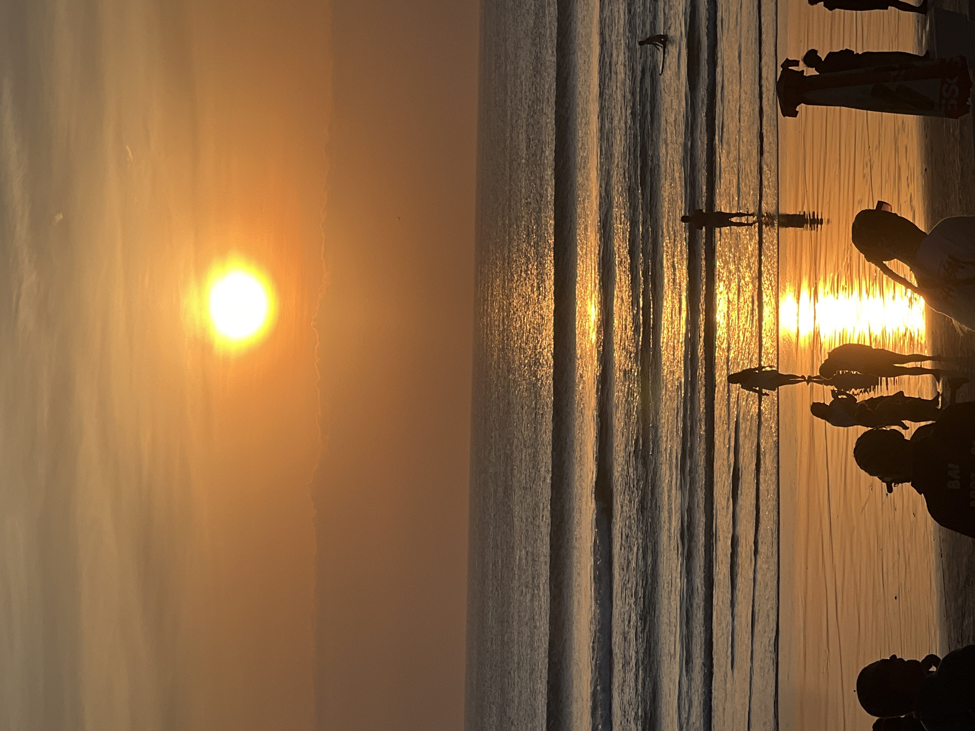 The image captures a serene sunset scene at Kuta Beach, Indonesia, with the sky painted in warm hues of orange and yellow. The sun, a bright orb, is positioned near the center of the frame, casting a shimmering reflection on the calm ocean. Gentle waves roll towards the shore, where several individuals are visible. In the distance, a lone surfer rides a wave. Closer to the beach, a few people stand in the shallow water, their silhouettes clearly defined against the vibrant sunset. On the wet sand, more figures are seen, some appearing to be walking or observing the sunset. The overall atmosphere is tranquil and picturesque, characteristic of a beach at dusk.