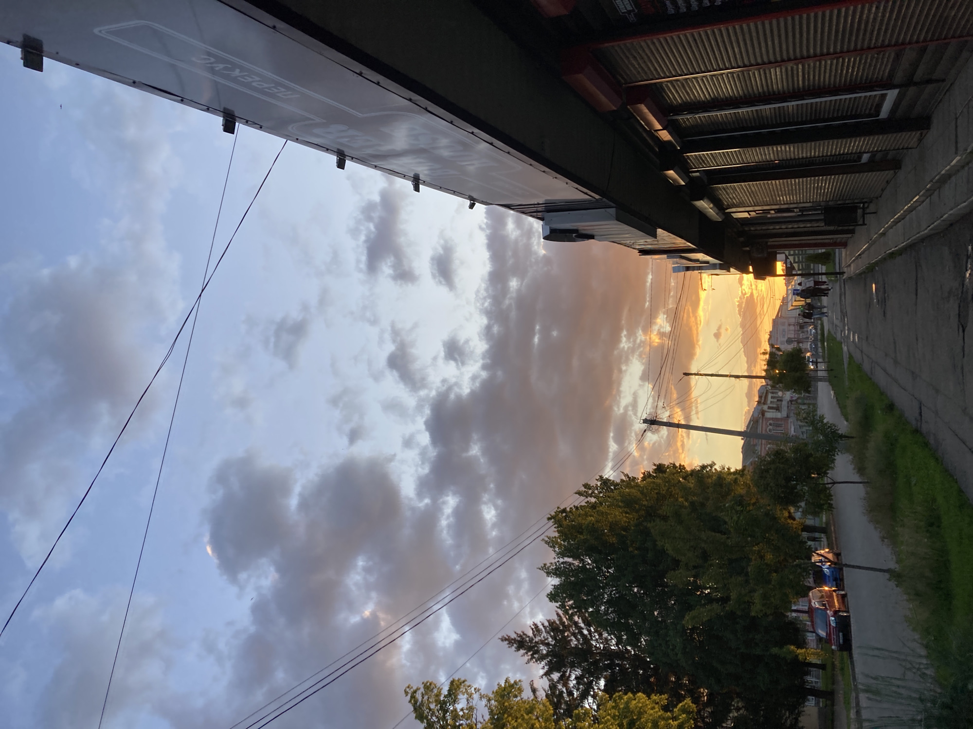 The image captures an atmospheric street scene at either sunrise or sunset, characterized by dynamic, warm lighting. The sky dominates the upper and left portions of the frame, transitioning from a pale blue and grey overhead to a vibrant golden orange and yellow on the right horizon, where the sun is setting or rising. Scattered clouds are illuminated by this warm light, creating an expressive canopy. Several thin, dark utility wires crisscross the upper sky, and a small bird is visible in flight in the upper left.
On the left side, a large sign is attached to a building, displaying white Cyrillic text. The words "СУПЕРМАРКЕТ" (SUPERMARKET) and "ПЕРЕКУС" (SNACK) are clearly visible, indicating a commercial establishment. Below this building, extending towards the mid-right, are the undersides of multiple structures with corrugated metal roofs, appearing as awnings or stalls, creating strong shadows and linear architectural patterns.
On the right side of the image, a paved street or sidewalk stretches into the distance, lined by lush green trees. Several cars are parked along the street beneath the trees. A utility pole stands prominently among the trees. In the far distance, silhouetted against the bright golden sky, are buildings with distinct architectural features and a few small figures, likely people, on the street. The overall scene conveys a quiet, reflective moment in an urban or suburban environment, with the Cyrillic text and architecture consistent with Poltava, Ukraine.