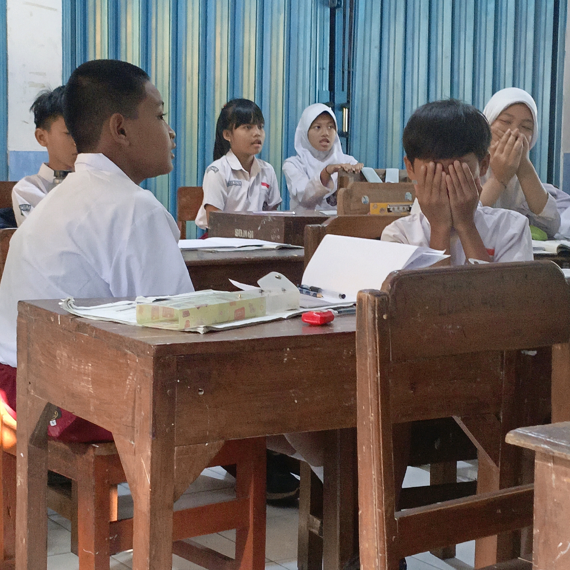 In Salatiga, Indonesia, this image captures a classroom scene with students in white uniforms sitting at wooden desks. In the foreground, a boy in a white shirt is partially obscured, with his hands covering his face, suggesting shyness or distress. To his right, a girl in a white hijab covers her mouth with her hands, her eyes visible and conveying a similar emotion. Further back, other students are seated, with one girl wearing a white uniform and a red and white badge, looking forward. The room is lit by overhead light, and a blue and white striped curtain is visible in the background. The desks are made of dark wood, and some have papers and pencil cases on them. The overall atmosphere appears to be one of a typical school day, with a focus on the students' reactions.