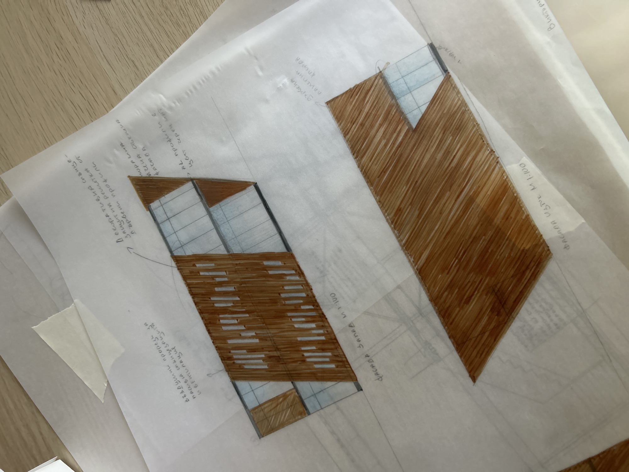 The image depicts architectural sketches on translucent tracing paper. Two geometric designs are visible, featuring brown-shaded sections with wood-like texture and blue-shaded areas possibly representing glass. Handwritten notes in Cyrillic are scattered around the drawings, including terms like "facade west M 1:100" and "facade east M 1:100." Other notations suggest "passive sun protection and ventilation," a "decorative sun-protective lattice of wooden profiles," and "panoramic glass." While the exact location is not explicitly stated, the sketches are likely related to a project in Sofia, Bulgaria, based on the Cyrillic language and the mention of "Viktor" with "Achena Staklena." The sketches appear to be preliminary designs or concepts for a building's facade. The presence of a wood table in the background suggests an office or studio environment.
