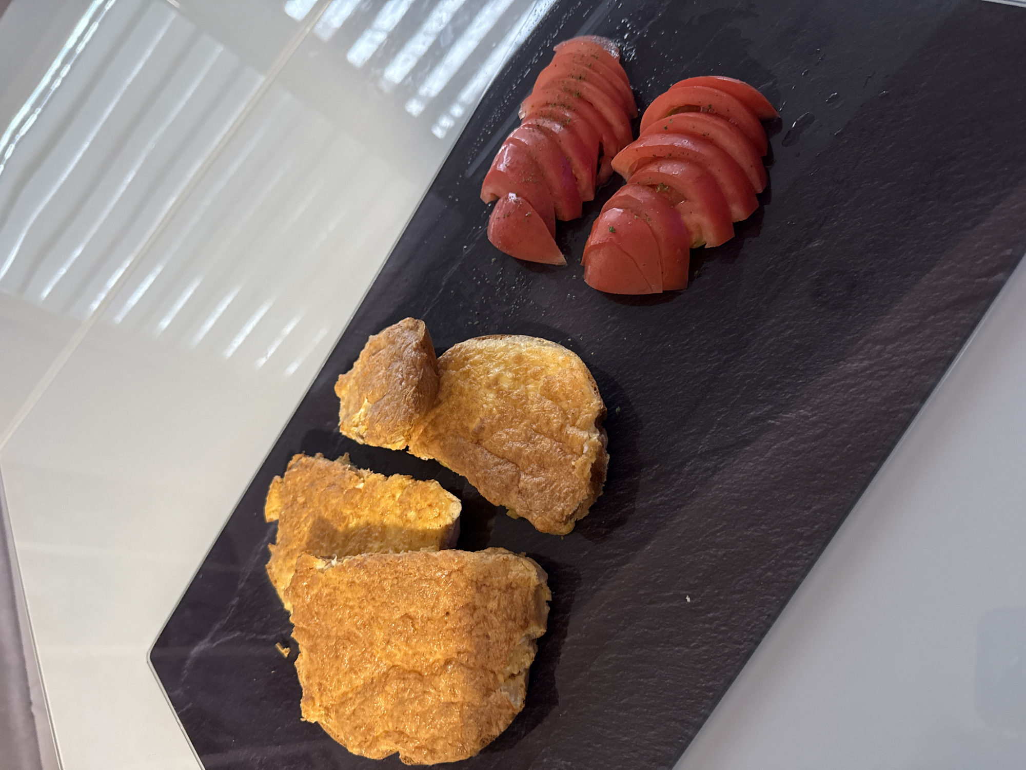 A close-up shows a black rectangular plate on a white tiled surface, possibly a countertop. The plate holds two distinct food items. On the upper right side are two piles of thinly sliced tomatoes, arranged overlapping each other, seasoned with green herbs. On the lower left side are three pieces of fried egg, each irregularly shaped, showing a golden-brown hue. The scene appears indoors. No people are visible. The location is undetermined.