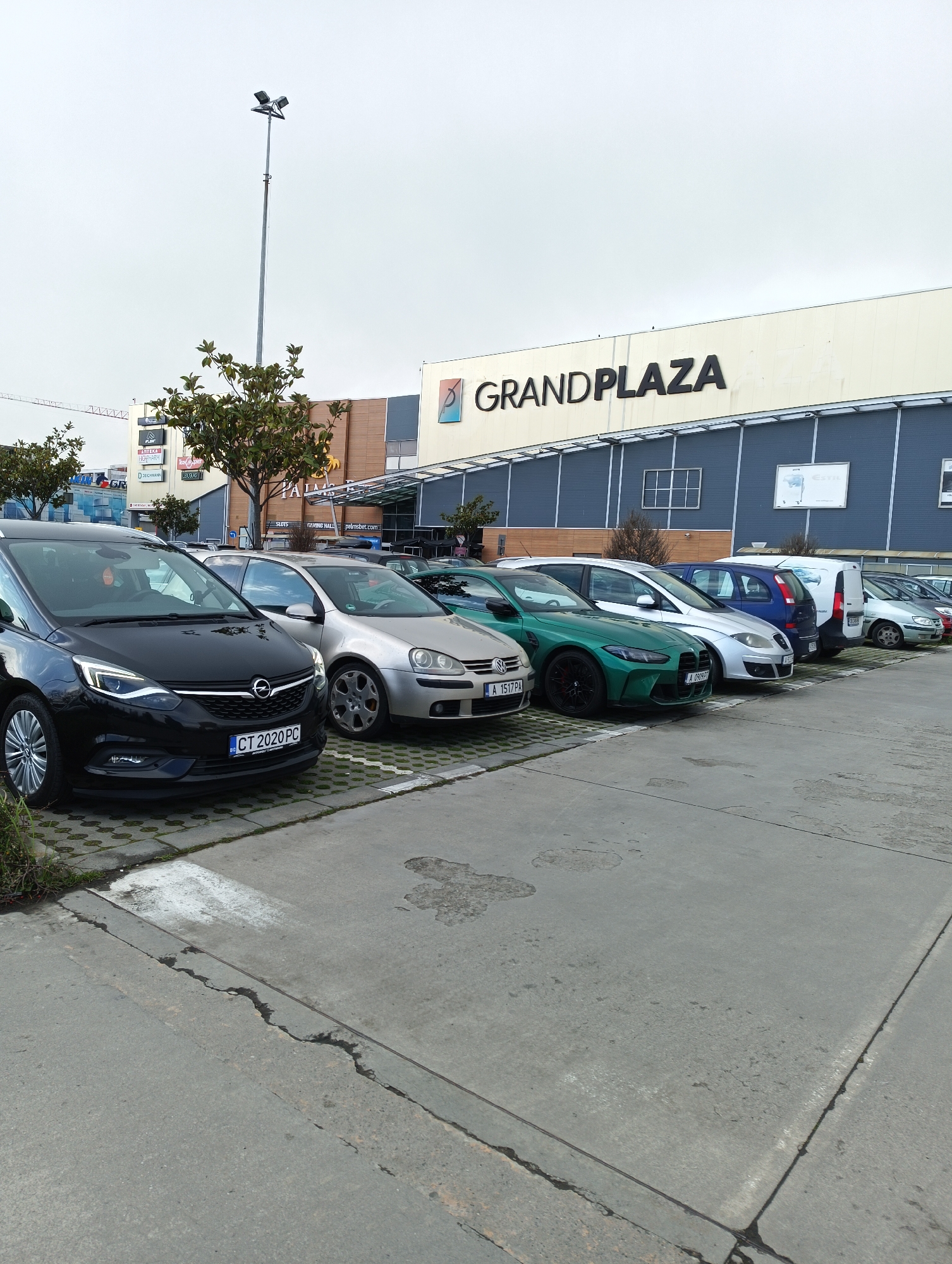 The image shows a parking lot in front of the Grand Plaza shopping mall. The location is likely in Burgas, Bulgaria, based on the appearance of Bulgarian license plates on the cars. There are several cars parked in a row including an Opel Zafira, Volkswagen Golf, and a green BMW. Behind the cars, the shopping mall is visible with the name "GRANDPLAZA" displayed on the facade. A tall light pole stands near the mall. The sky is overcast. There are also additional store signs visible, including HIGH-PHARM, DEICHMANN, and LOUGIAS. A sign for a gaming hall called "palmsbet.com" is also visible.