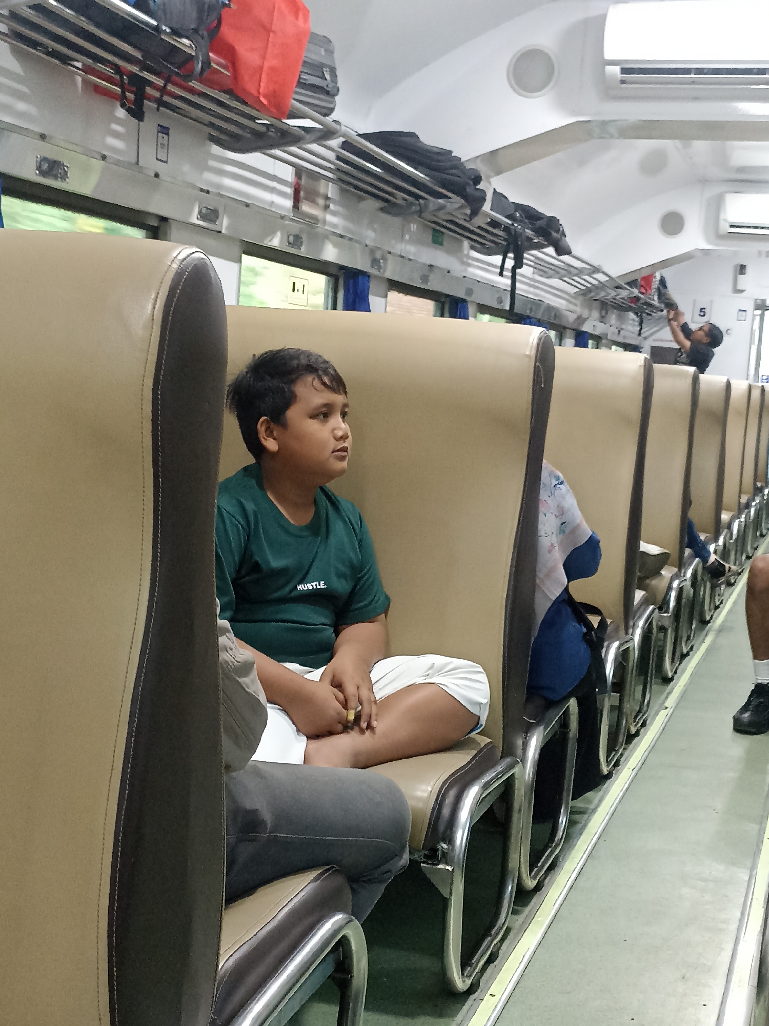 The image depicts the interior of a train carriage, likely in Rogojampi, Indonesia, showing a young boy sitting on a tan-colored seat, looking to the right. He is wearing a dark green t-shirt with the word "HUSTLE." printed on it, and white shorts. His hands are clasped in his lap. The seats are arranged in rows, with luggage stored on overhead racks. In the background, other passengers are visible, including a person in a blue headscarf and another person reaching for an item on the rack. The floor of the carriage appears to be light green, and the walls are white with overhead air conditioning units. The lighting suggests it is daytime.