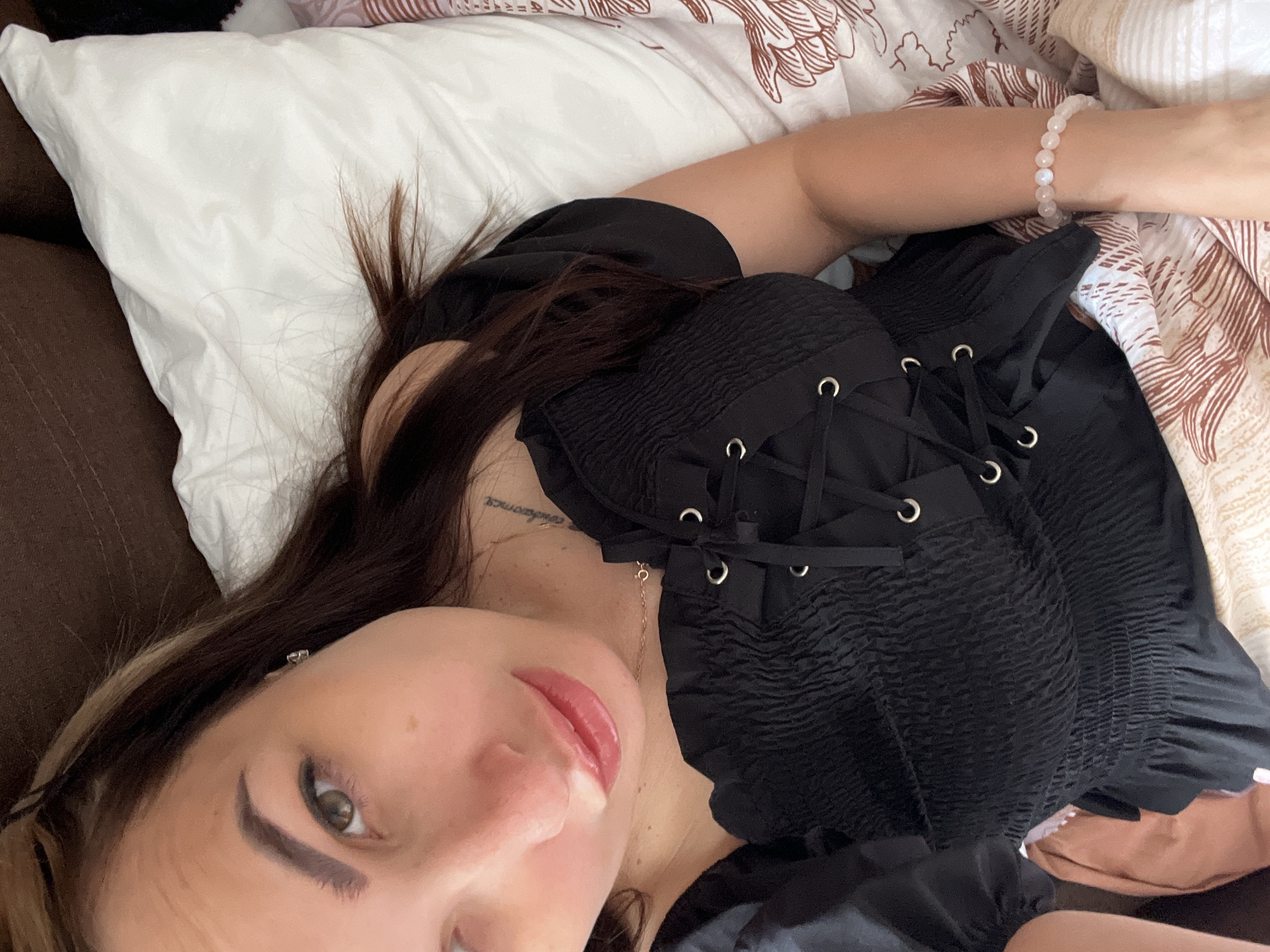 A close-up, high-angle shot shows a woman lying down, facing upwards, with her head resting on a white pillow. She has long, dark brown hair with some lighter strands mixed in, and her eyes are open and looking towards the camera. Her lips are slightly parted, and she is wearing a black, smocked top with a corset-style lace-up front. A delicate gold necklace is visible around her neck, and a tattoo can be seen on her upper chest. She is also wearing a bracelet made of light pink beads on her right wrist. The background consists of a brown couch and a patterned bedspread.
