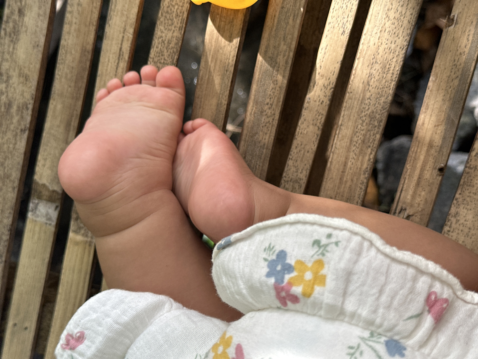 A high-angle shot shows the bare feet of a child, possibly an infant, resting on a bamboo or rattan-style wooden surface. The left foot is more prominent, displaying the sole and toes. The right foot is partially obscured behind it. The child is wearing a white cloth garment with a floral pattern, featuring flowers in blue, yellow, and pink. The background includes a glimpse of what appears to be a natural, possibly outdoor, environment, with light reflecting off a surface that could be water. The setting is implied to be in North Kuta, Indonesia.