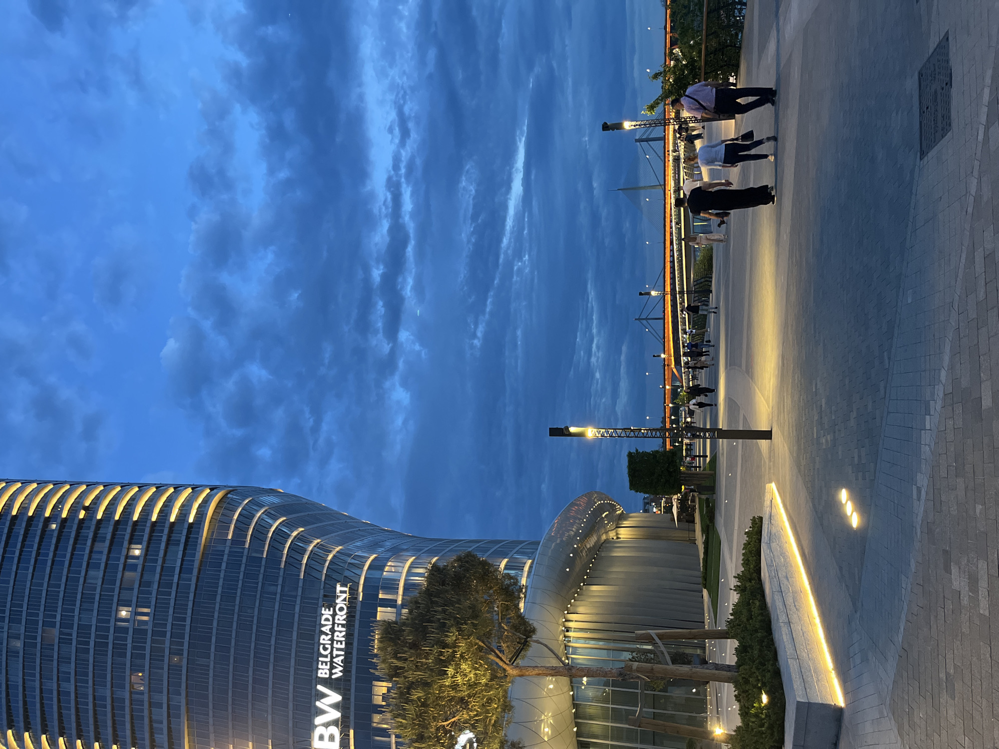 The image captures an evening scene at the Belgrade Waterfront in Serbia. A modern, curved skyscraper with illuminated bands and the "BW BELGRADE WATERFRONT" logo dominates the left side of the frame. In the foreground, a tree is partially lit. To the right of the building, a walkway is lined with illuminated elements and a short wall with shrubbery. People are walking along the walkway towards a bridge in the distance. The sky above is filled with clouds.