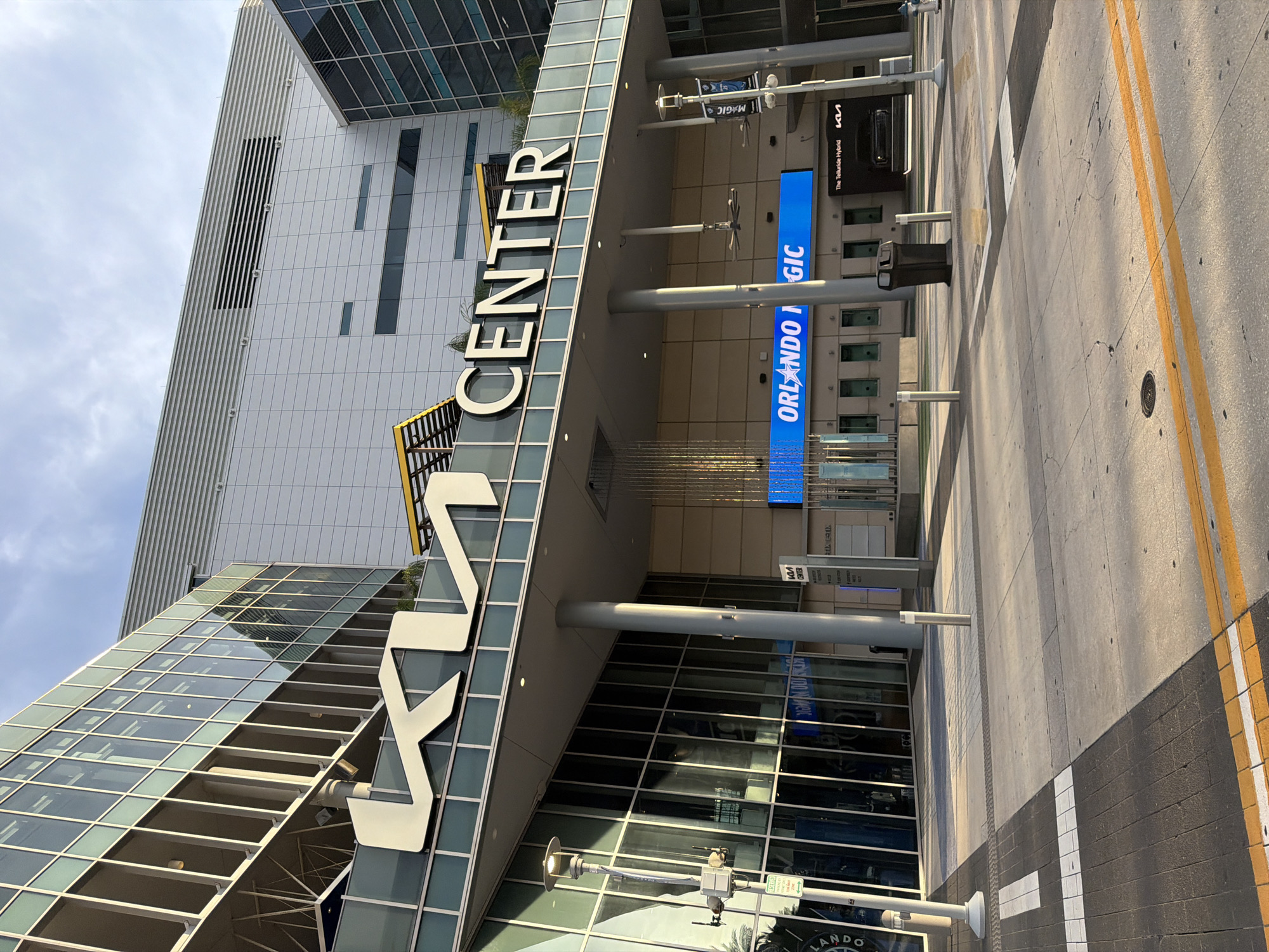 The image shows the exterior of the Kia Center, located in downtown Orlando, Florida, United States of America. The building features a modern architectural design with a glass facade and prominent signage. The large white "KIA" logo is prominently displayed above the entrance, followed by the word "CENTER" in white lettering. Below the signage, a large blue LED screen displays "ORLANDO MAGIC" with a star graphic. The area in front of the building is paved with concrete and includes street markings, bollards, and a trash receptacle. The scene is captured during daylight, with sunlight casting shadows on the ground, indicating a bright and clear sky. There are no people visible in the image. The overall impression is one of an active urban environment with a focus on the sports and entertainment venue.