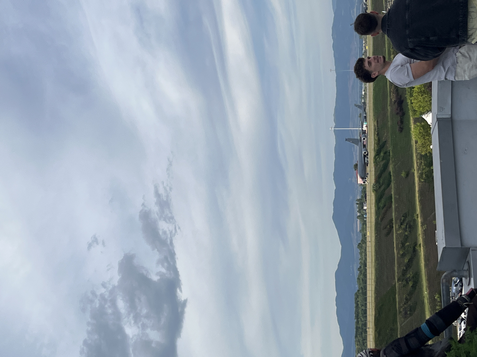 This image captures a view of an airfield with multiple planes on the tarmac. In the foreground, two men are standing on a rooftop, facing away from the camera and towards the airfield. One man wears a light grey t-shirt and light-colored shorts, while the other is dressed in a dark denim jacket. To the left, partially visible, is a person wearing motorcycle gear. The background features a landscape of green hills and mountains under a cloudy sky. The airfield appears to be located near a populated area, with some structures and vehicles visible. Based on the specific types of aircraft visible, which are likely military transport planes, and the geographical context of Sofia, Bulgaria, this scene is likely depicting the Sofia Airport, potentially a section used for cargo or military operations. The overall atmosphere is overcast and somewhat industrial, contrasting with the natural landscape in the distance.