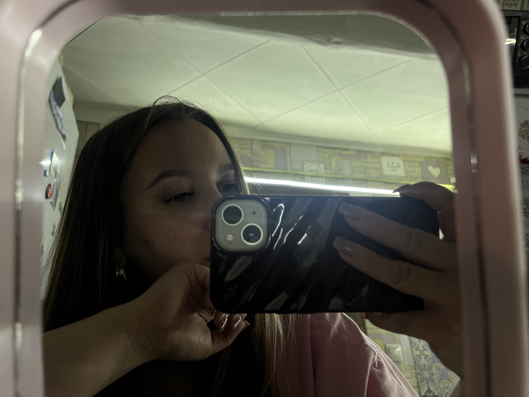 A female individual is taking a selfie using a smartphone in front of a mirror. The mirror has a light pink frame. The individual is holding a black smartphone with a dual-lens camera visible on the back. The person's face is partially visible, with dark hair, fair skin, and an earring. Her right hand is touching her face, and her left hand is holding the phone. She's wearing a light pink shirt. The reflection in the mirror shows a room with a tile-patterned wall and a light fixture near the ceiling. A shelf and other items are visible in the background to the upper right. The location is presumed to be indoors, possibly a bathroom. There is no indication of the location in the city of Port-Katon, Russia.