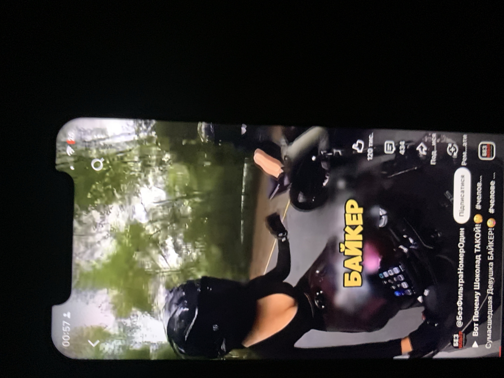 The image shows a vertical video displayed on a smartphone screen. The video features a person wearing a black helmet and a black outfit, riding a motorcycle. The text overlay on the video reads "БАЙКЕР" in yellow letters. Below this, there is an account handle "@БезФильтраНомерОдин" and a "Подписаться" button. Further text indicates a title "Вот Почему Шоколад ТАКОЙ!" and "Сумасшедшая Девушка БАЙКЕР!". Interaction icons for likes, comments, and shares are visible on the right side of the screen. The background of the video appears to be a road with trees. There is no indication that this scene is located in Zhmerynka, Ukraine.