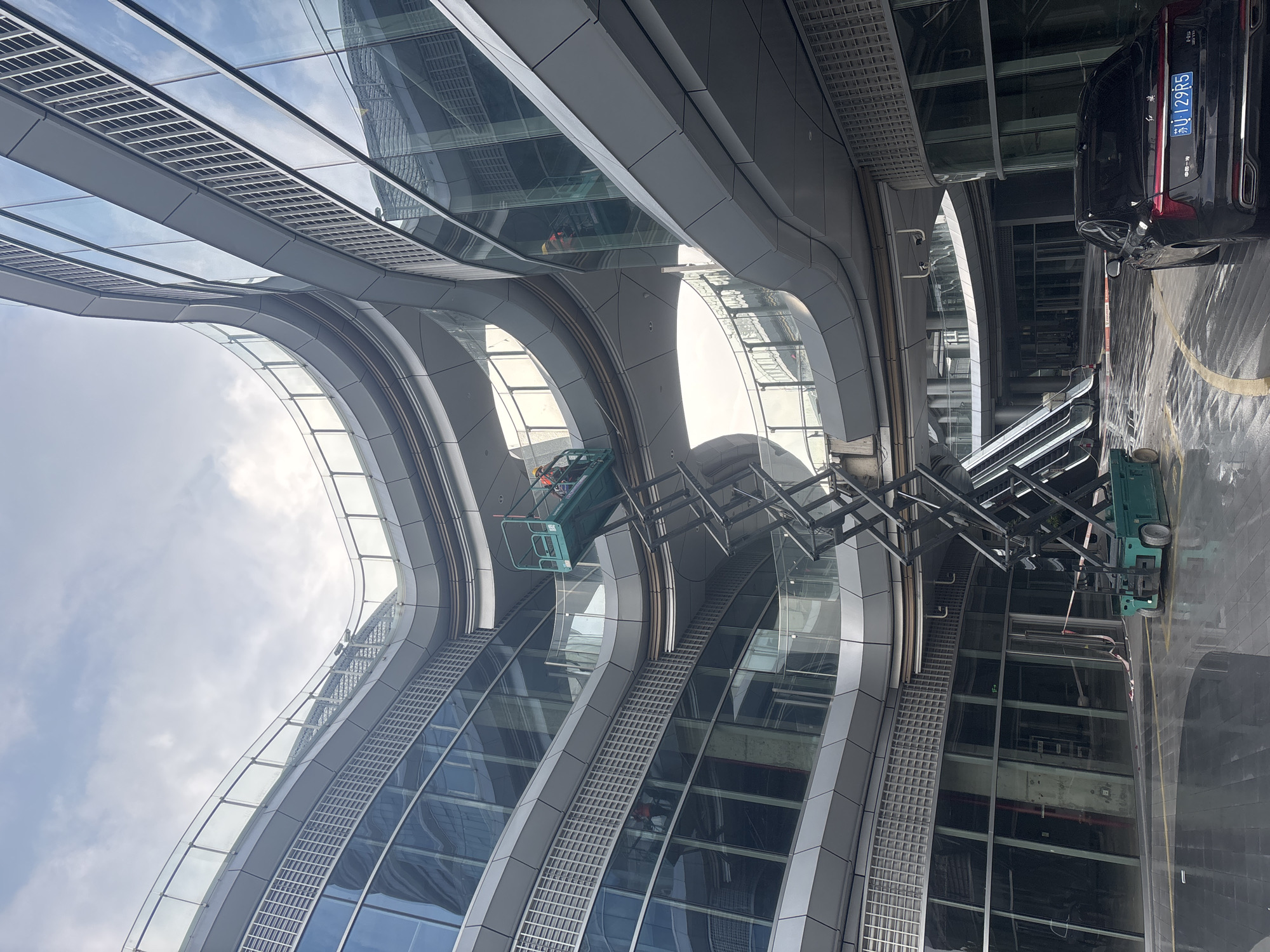 This is a photograph of the exterior of a modern, multi-story building with curved architectural elements and a facade of glass and metal. The building appears to be under construction or undergoing maintenance, as indicated by a green scissor lift with two construction workers in hard hats operating at mid-level. One worker is visible on the platform of the lift. In the foreground, a black sedan is parked on a wet, paved surface with yellow lane markings. The sky above is overcast with scattered clouds. The scene is located in the Suzhou Industrial Park, China.