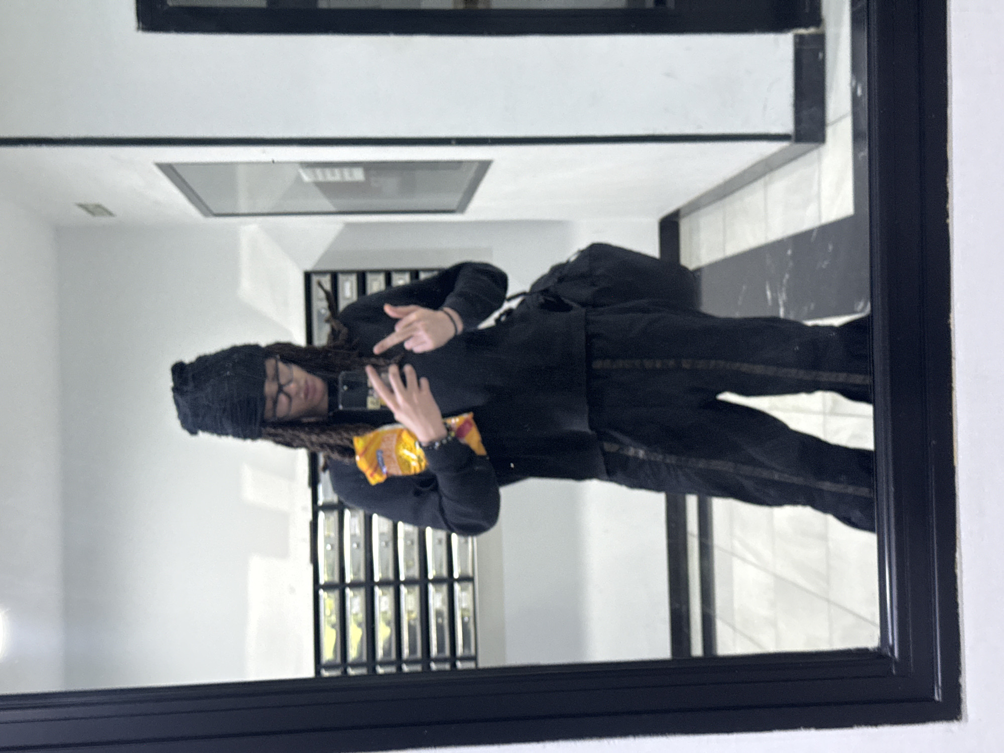The image depicts a mirror selfie of a person in an indoor setting. The individual, appearing to be male, has dreadlocks and is wearing glasses, a black beanie, and an all-black outfit consisting of a hoodie or jacket and pants. A black backpack is visible on their back.
The person is holding a smartphone in one hand, presumably to take the photo, and in the other hand, they are holding a yellow bag of snacks, possibly chips, with some illegible text on the packaging. They are making a defiant or casual hand gesture, appearing to be a middle finger, with the hand not holding the phone.
The background, seen as a reflection in the mirror, shows a brightly lit interior space. The walls are predominantly white, complemented by architectural details with black frames or outlines. The floor has a black and white tiled or patterned design. To the left side of the reflection, a series of vertically stacked, dark-framed compartments resembling mailboxes or storage lockers are visible. A rectangular light fixture is reflected on the ceiling. The overall impression is that of a modern building's common area or hallway.
The time of day is not discernible due to the indoor setting, and no specific external weather cues are present. There are no direct visual cues within the image to confirm the provided location context of Madrid, Spain, beyond the building's contemporary design.