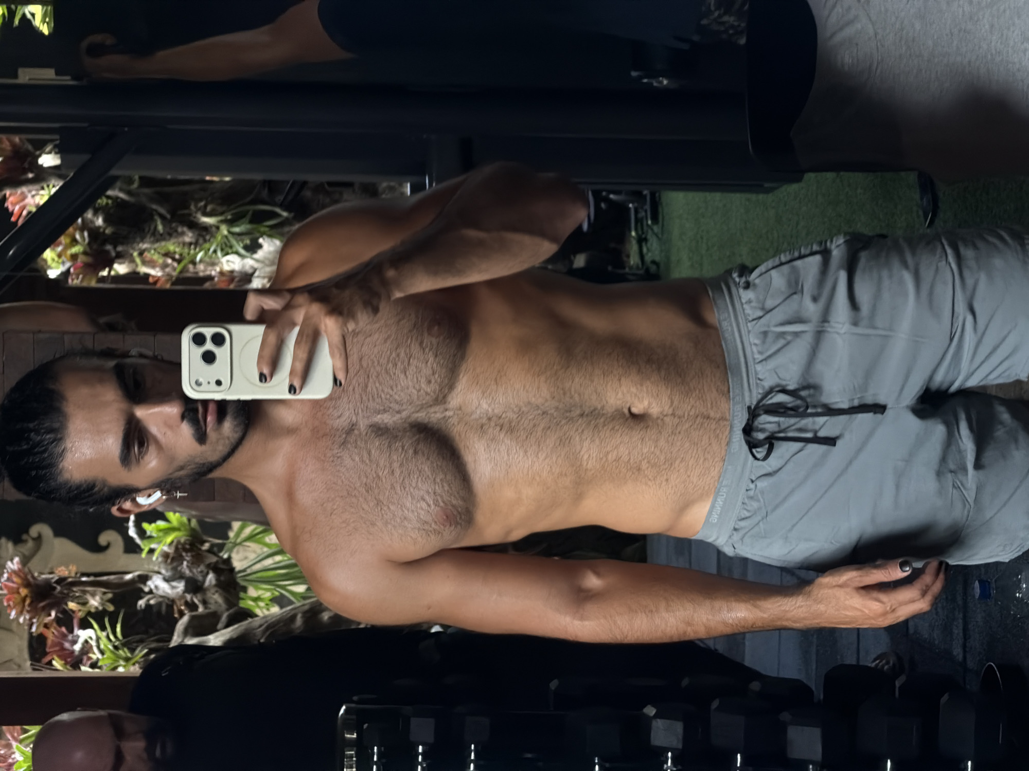 A man with dark hair pulled back in a bun is taking a mirror selfie in a gym. He is shirtless, wearing grey athletic shorts, and has dark nail polish on his fingernails. He is also wearing an earbud in his left ear with a cross-shaped earring. To his left, another man is partially visible, wearing a black shirt. The background of the gym includes plants, mirrors, and weight racks with dumbbells. The location is specified as North Kuta, Indonesia.