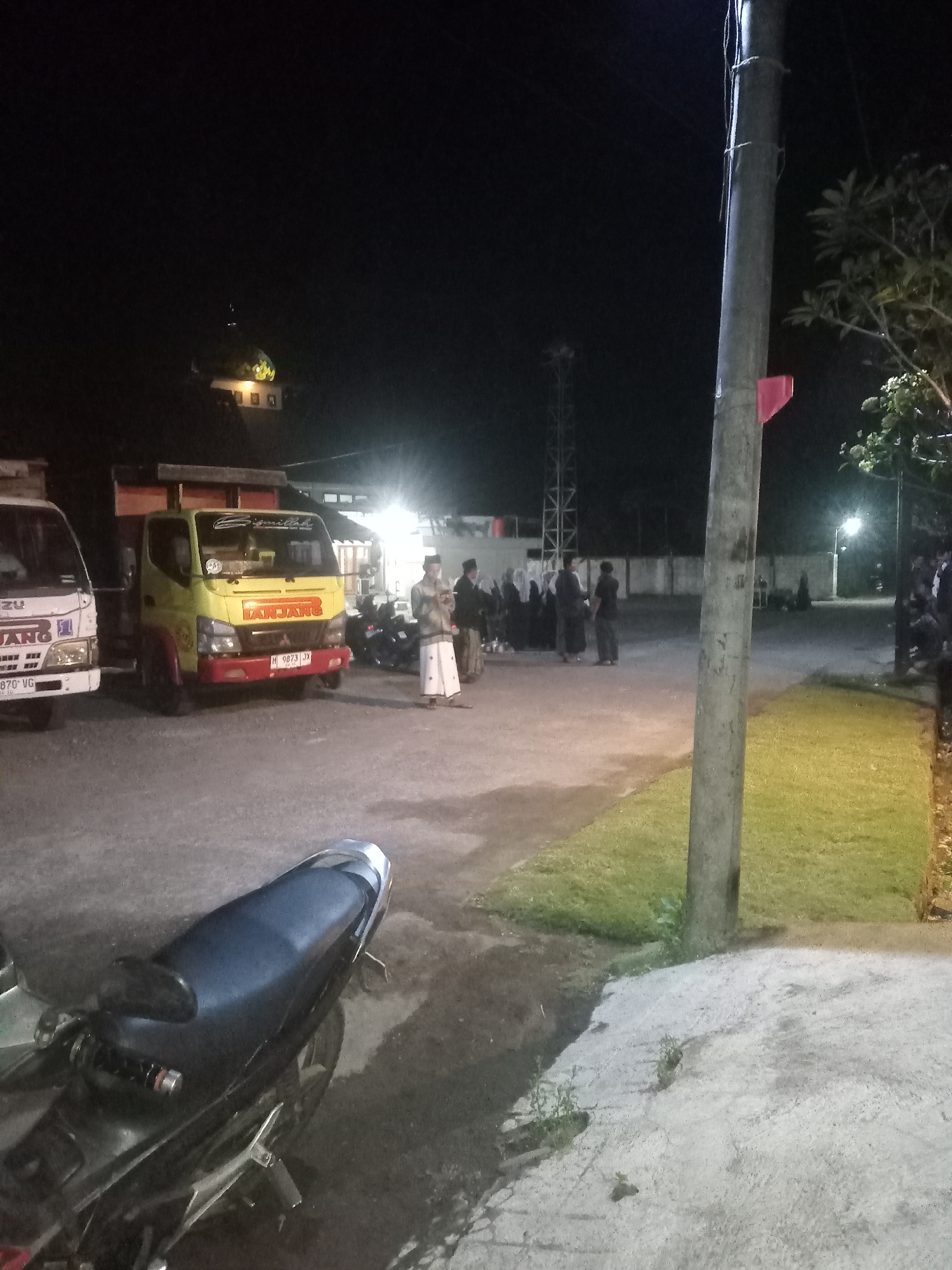At night in Majaksingi, Indonesia, a group of people are gathered near several vehicles, including a white Isuzu truck and a yellow Mitsubishi Fuso truck. The yellow truck has the word "TANJANG" visible on its side. Several motorcycles are parked behind the trucks. In the background, a mosque with a green dome is illuminated. The scene is lit by artificial lights, casting shadows and highlighting the vehicles and people. A prominent utility pole stands in the foreground, to the right of the frame. The ground appears to be a mixture of asphalt and concrete. The overall atmosphere is one of a community gathering or event taking place after dark.