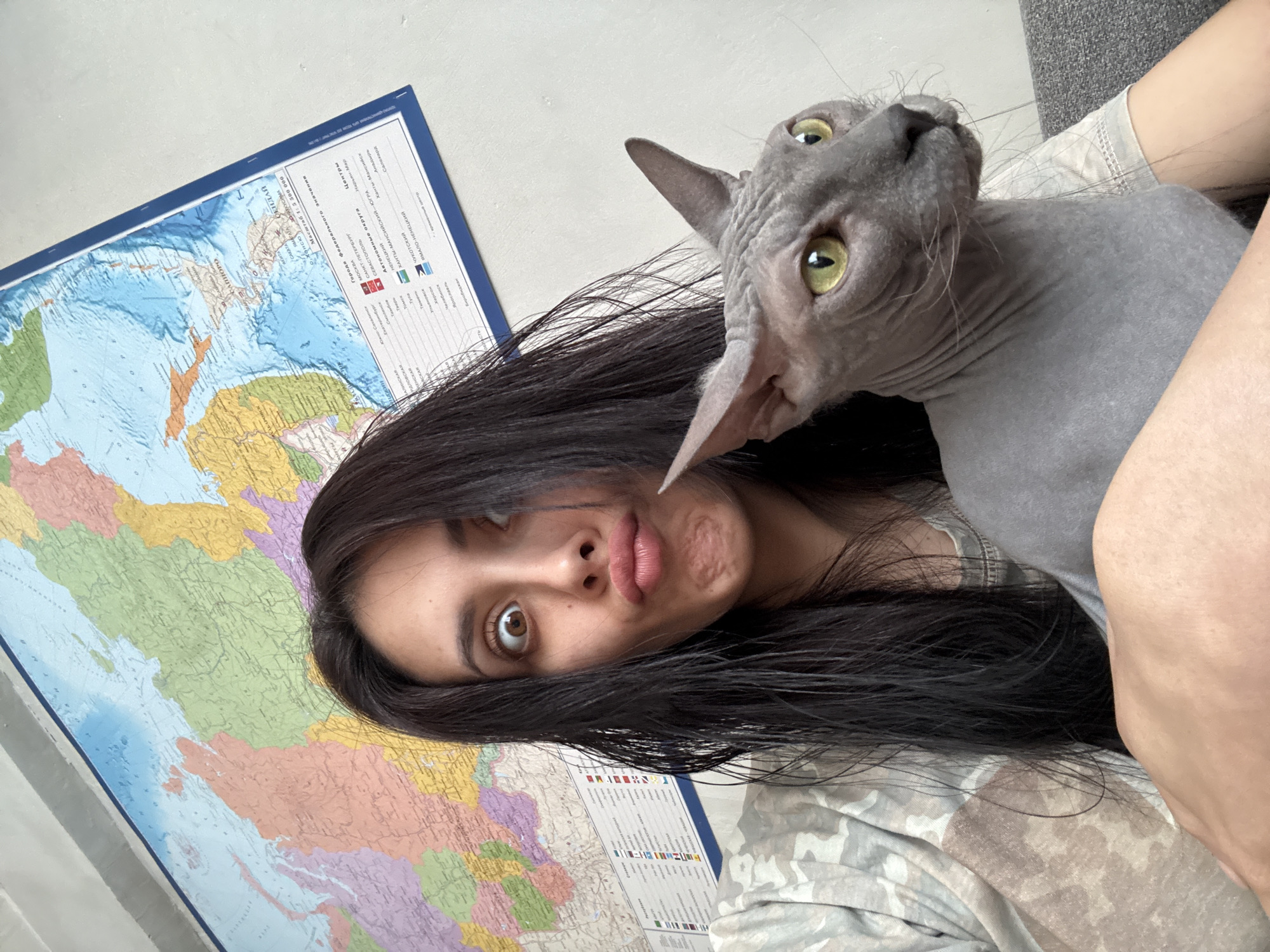 A photograph features a young woman holding a Sphynx cat, with a political map of the world visible in the background. The woman has long, dark hair and is making a duck face. The hairless cat is gray with yellow eyes and looking upwards. The map is colorful and includes details such as cities, borders, and different geographical regions. The photo appears to be taken indoors, as suggested by the plain wall and the edge of a gray couch in the upper right corner. The location of the image in the city of Votkinsk, Russia, is not discernable from the content provided.