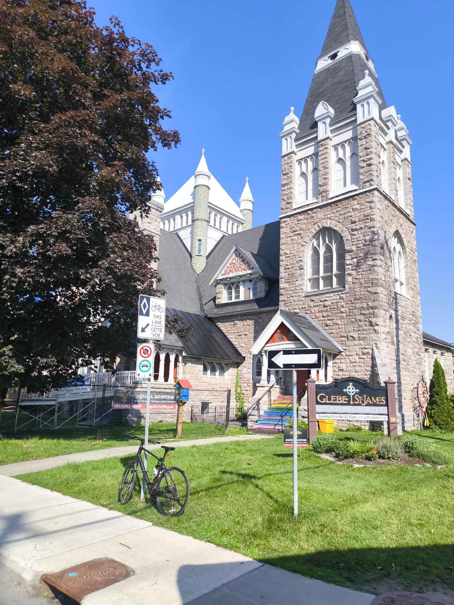 The image shows the exterior of Glebe St. James United Church, located in the Glebe neighborhood of Ottawa, Canada. The church is a stone building with a tall, dark-sloped steeple. The entrance features rainbow-colored steps leading up to a red door. In front of the church, a sign reads "GLEBE ST. JAMES UNITED CHURCH." A black bicycle is parked on the grass near a sidewalk. Various traffic and informational signs are also visible, including one indicating a bike lane and another with a prohibition symbol. The scene is set against a clear blue sky, with a large tree to the left and green grass and landscaping around the church.
