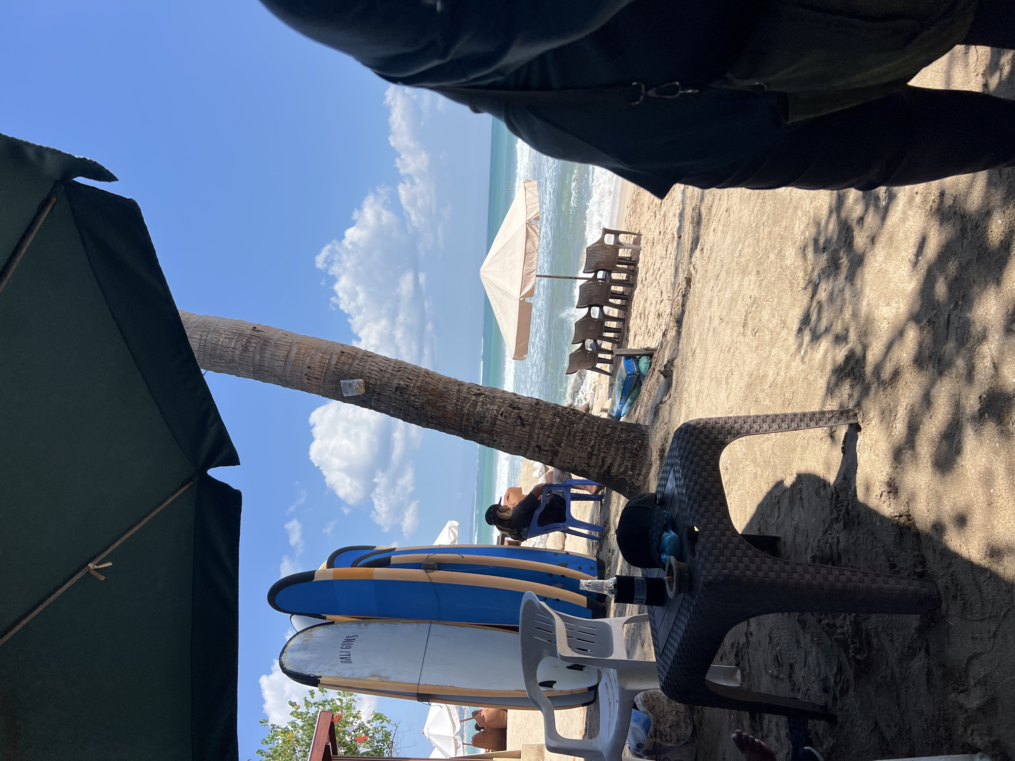 The image depicts a beach scene in Kuta, Indonesia, under a bright, sunny sky with scattered clouds. In the foreground, a dark brown, woven plastic table sits on the sand, with a bottle in a cooler, a black baseball cap, and a small ashtray on its surface. To the left of the table, a white plastic chair is visible, with another person's feet partially in frame. Behind the table, several surfboards are propped up, two of which are blue and white, with text that appears to read "BALI GUNS" on one. A person sits in a blue plastic chair further back, facing the ocean, dressed in dark clothing and a baseball cap. To the right of the scene, a palm tree trunk is prominent, and further in the distance, beach chairs and a beige umbrella are set up on the sand next to the turquoise ocean with moderate waves. The overall atmosphere suggests a casual beachside establishment.