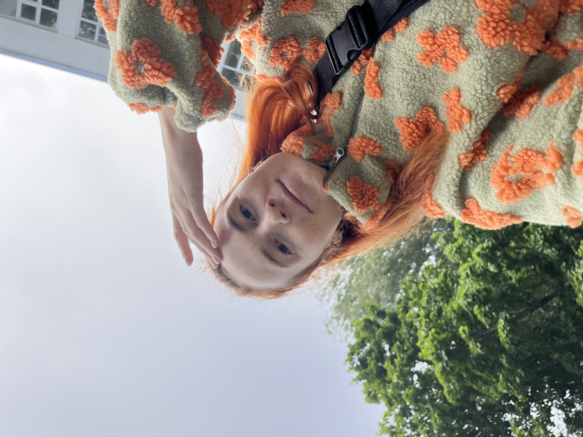 A woman with bright orange hair is taking a selfie outdoors on an overcast day. She is wearing a light green fuzzy jacket with an orange floral pattern and a black crossbody bag strap visible. Her left hand is raised to her forehead. In the background, lush green trees are visible to the left, and a white building with windows can be seen to the right. The overall setting suggests an outdoor urban or suburban environment. The specific location in the city of Chaikovsky, Russia, cannot be determined from the visual information in the image.