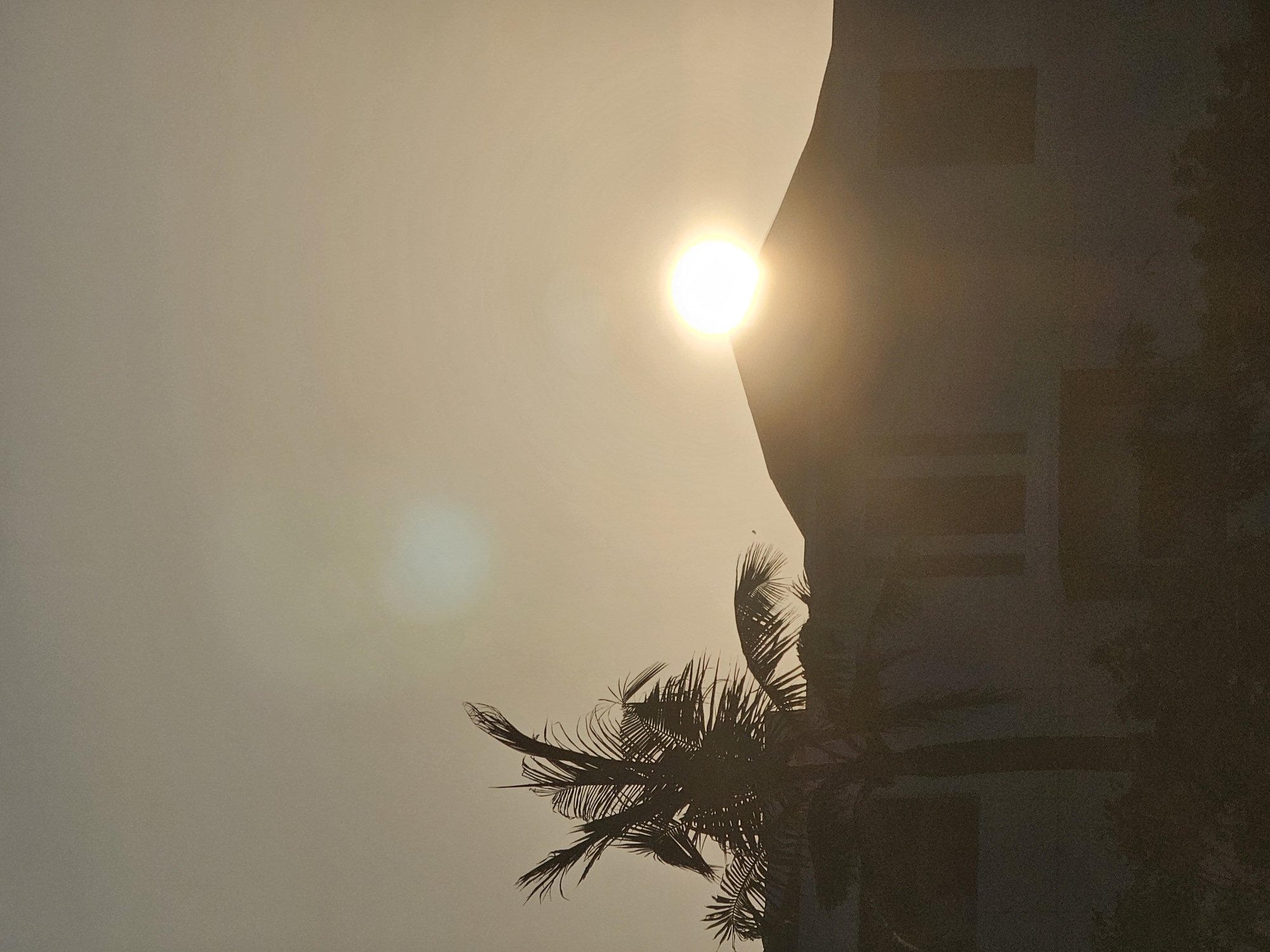 A low-angle vertical shot captures the sun setting behind a silhouetted building and a palm tree. The sun is a bright orb, partially obscured by the roofline of the building. The sky is a pale orange, with a lens flare visible as a smaller, bright spot in the upper portion of the frame. The palm tree, on the left side of the image, is also silhouetted against the sky, its fronds appearing wispy and delicate. The building appears to be a multi-story structure with dark rectangular windows. The overall impression is one of a peaceful, tropical evening. The location is the city of Denpasar, Indonesia.