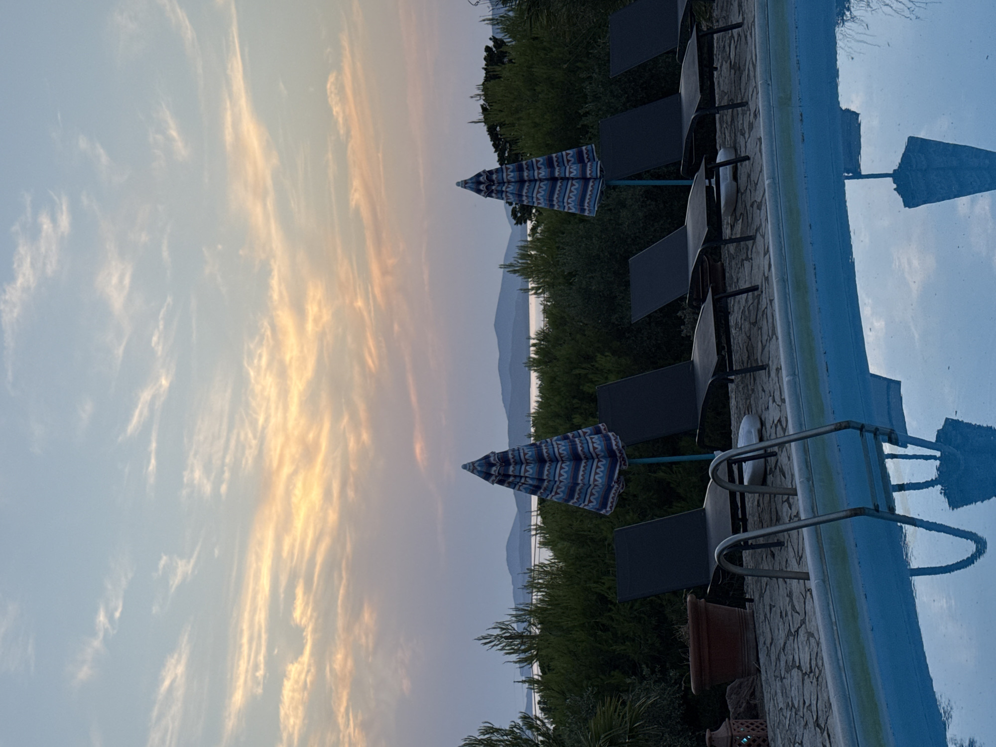 This image depicts an outdoor swimming pool area at dusk, with a body of water and distant hills visible in the background. The sky is filled with wispy clouds illuminated by the setting sun, casting warm hues of orange and pink. Several empty lounge chairs are arranged along the poolside, some with umbrellas partially open. A terra cotta pot and a decorative lantern are visible on the stone-paved area behind the chairs. The pool water is a vibrant blue, reflecting the sky and some of the surrounding elements. The overall scene suggests a tranquil, resort-like setting, possibly located in a coastal area near Alghero, Italy, given the presence of water and hilly terrain suggestive of the Sardinian landscape. No people are present in the image.