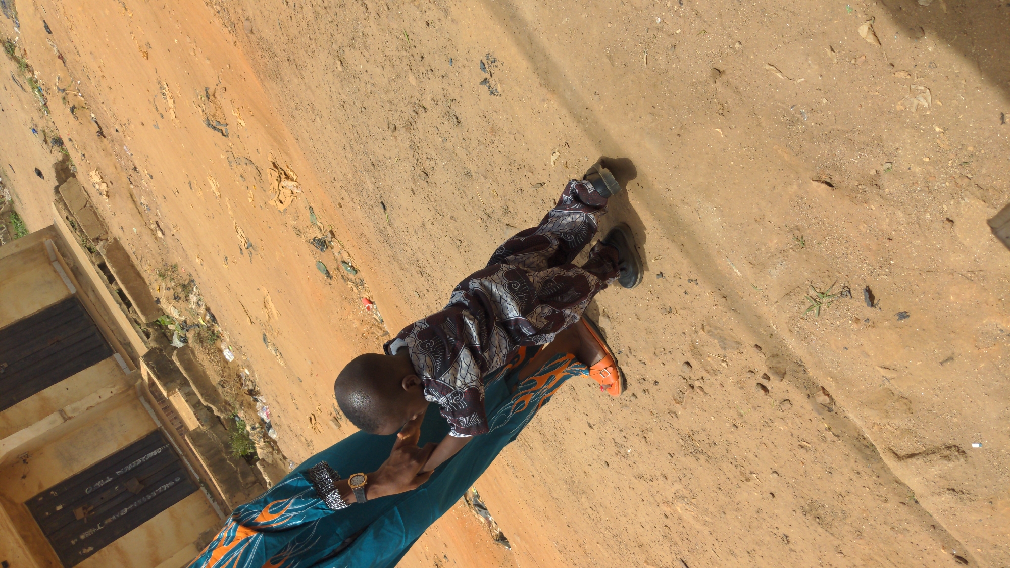 A high-angle shot shows a young boy carried on the hip of an adult. The boy wears patterned clothing, and shoes are on his feet. The adult wears a teal-colored outfit with orange patterns and orange sandals. The adult wears a watch on the wrist. The scene appears to be Ado, Nigeria. The background shows a dirt road and part of a building. Debris and small vegetation appear in the dirt.