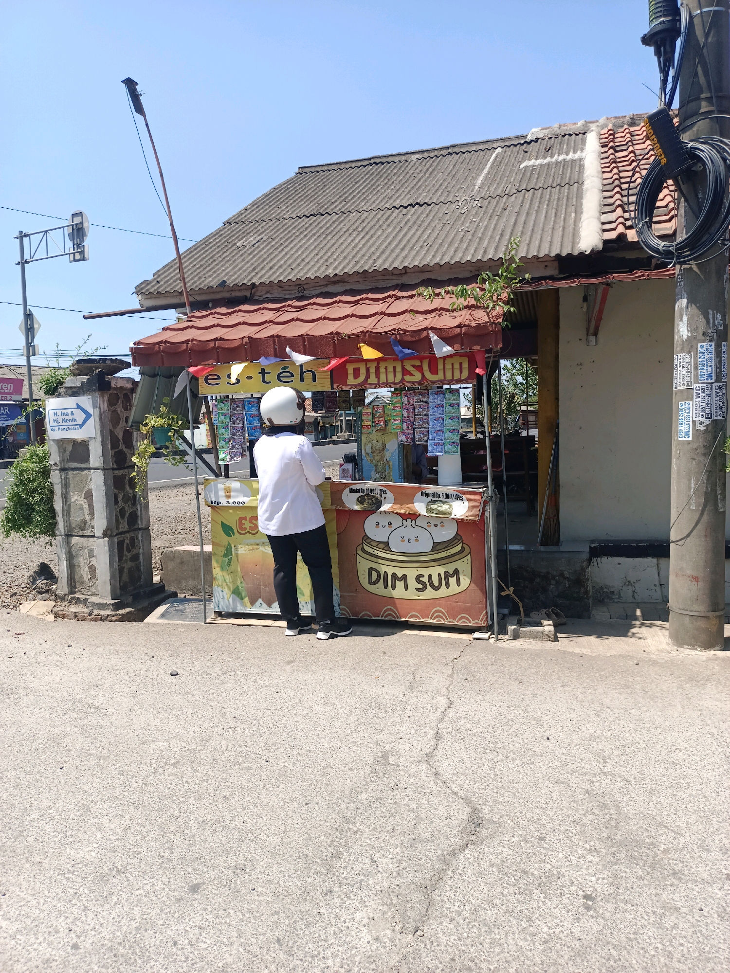 A person wearing a white shirt, black pants, and a white helmet stands at a street-side stall in Subang, Indonesia. The stall, covered by a red awning, displays colorful flags and sells "Dim Sum" and "Es Teh" (iced tea). The background shows a building with a corrugated roof and a utility pole with cables. The street in front of the stall is asphalt. A signpost with directions is visible to the left of the stall. The sky is clear and blue.