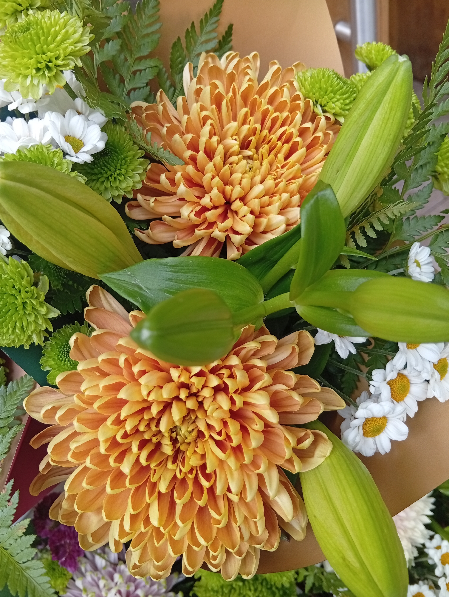 A close-up photograph shows a bouquet of flowers. The predominant flowers are two large orange chrysanthemums with tightly packed petals. Interspersed are green chrysanthemums, white daisies with yellow centers, fern fronds, and several unopened lily buds, recognizable by their smooth, green casings. The arrangement appears to be freshly assembled, with a mix of textures and colors. A light brown wrapping material is partially visible, suggesting the flowers are arranged in a bouquet. The specific location within Buenos Aires, Argentina, cannot be determined from the visual information provided. There are no people visible in the image.