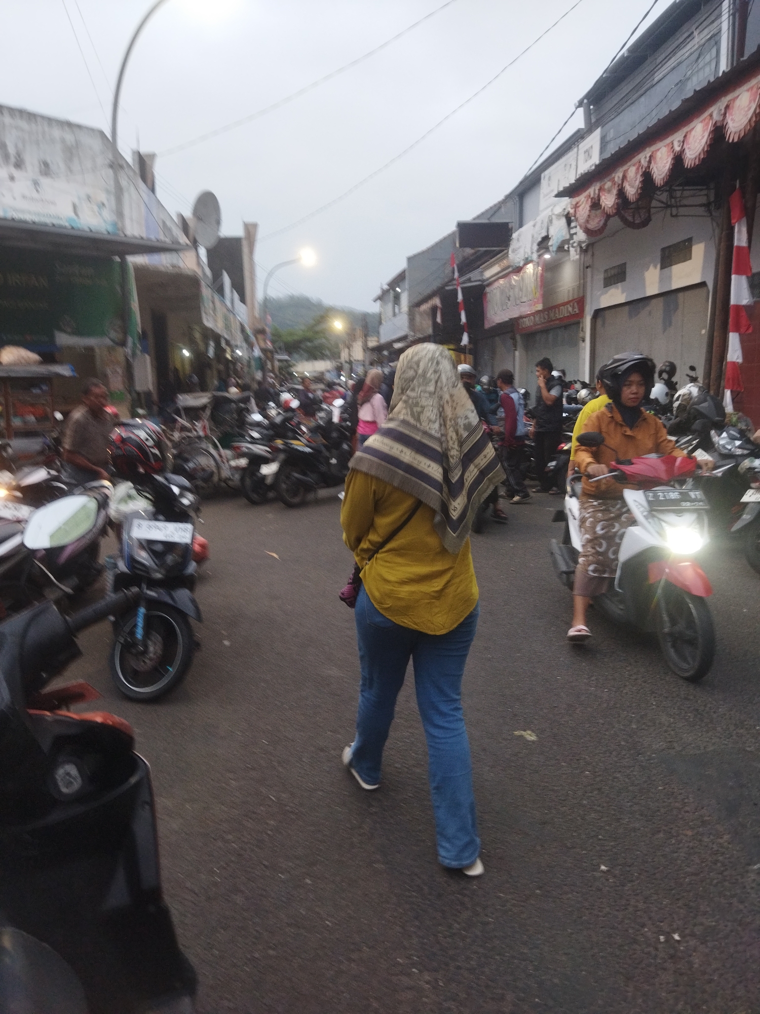 The image depicts a street scene in Banjar, Indonesia, likely a market or commercial area, with a cloudy sky. In the foreground, a person wearing a yellow top and blue jeans walks away from the camera, with a patterned headscarf. To the right, a person on a white and red scooter, wearing a brown jacket and patterned pants, rides towards the camera. The street is lined with parked motorcycles and appears to be a busy thoroughfare. Buildings with shops and awnings are visible on either side of the street. The overall lighting suggests it is either early morning or late afternoon.