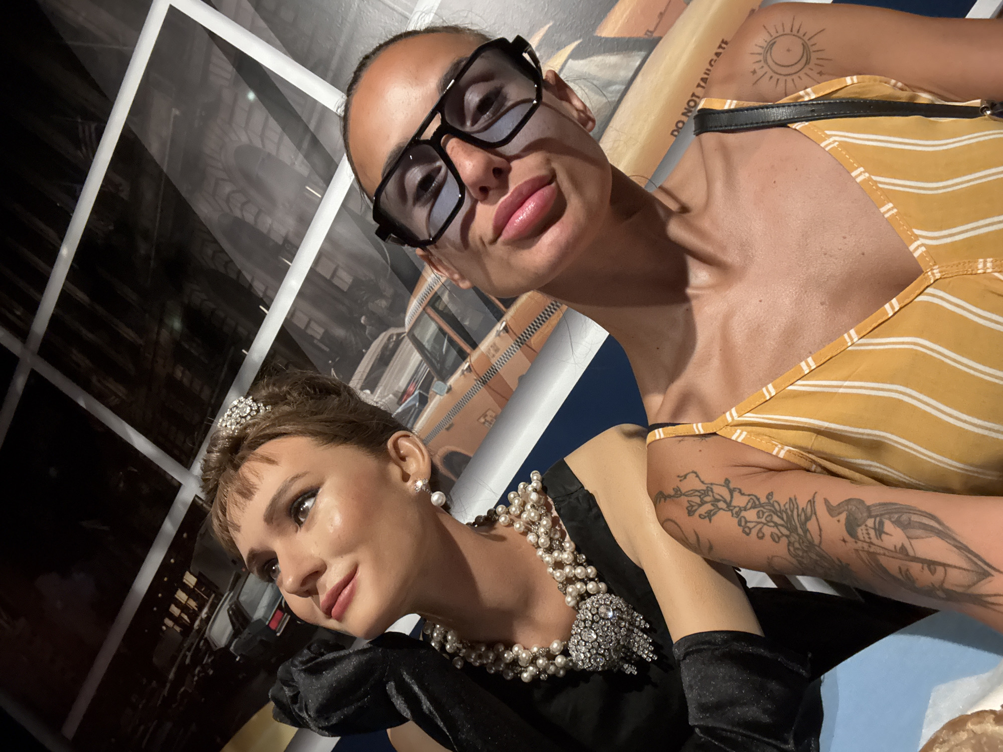 A woman with dark hair and sunglasses takes a selfie with a wax figure of Audrey Hepburn, who is dressed in a black dress, pearls, and gloves. The wax figure is positioned to the left of the woman, looking upwards and to the left. The background appears to be a display depicting a street scene, possibly in New York City, with a yellow taxi and buildings. The woman is wearing a yellow and white striped top and has tattoos on her right arm. The scene takes place indoors, likely in a museum or wax exhibit. There is no indication of Prague, Czechia in the image.