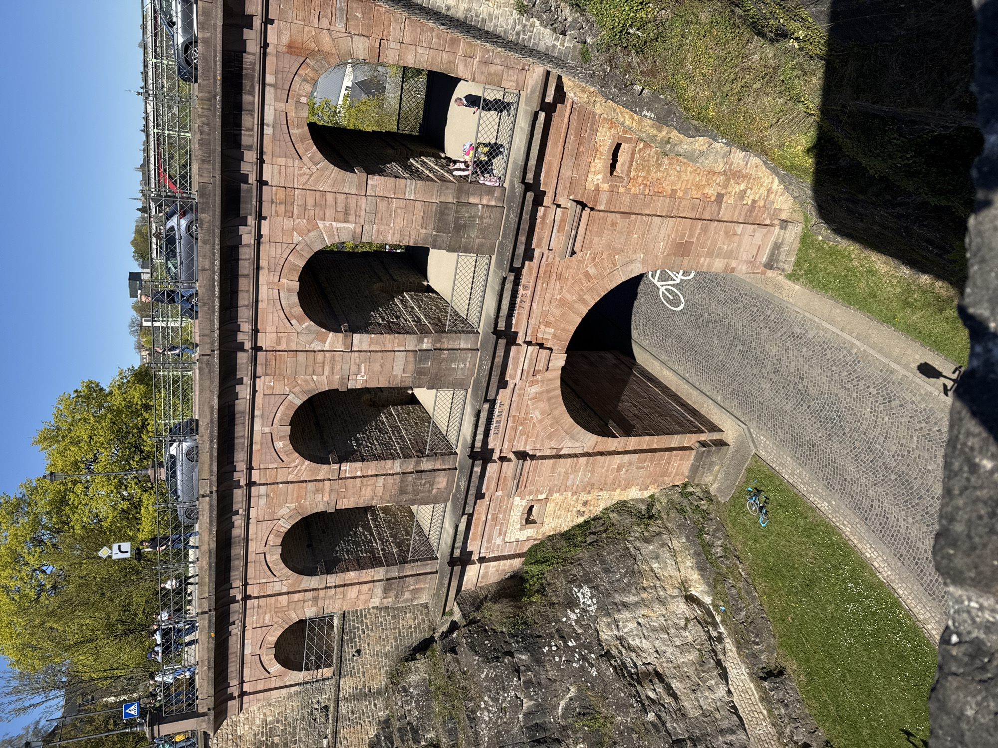 The image shows a section of the Passerelle, a large, arched stone bridge located in Luxembourg City, Luxembourg. The bridge features multiple arches that allow passage underneath. On top of the bridge, people and cars can be seen moving along the pedestrian and road lanes. Below the bridge, there are walkways with wrought iron railings, and one of the arches leads to a paved area marked with a bicycle symbol, indicating a bike path. A parked bicycle sits on the grass next to the bike path. The bridge's stone construction shows signs of weathering and age. The adjacent area is a mix of rocky terrain, vegetation, and cobblestone pathways. The angle suggests the photo was taken from an elevated vantage point, possibly another part of the bridge or a nearby structure.