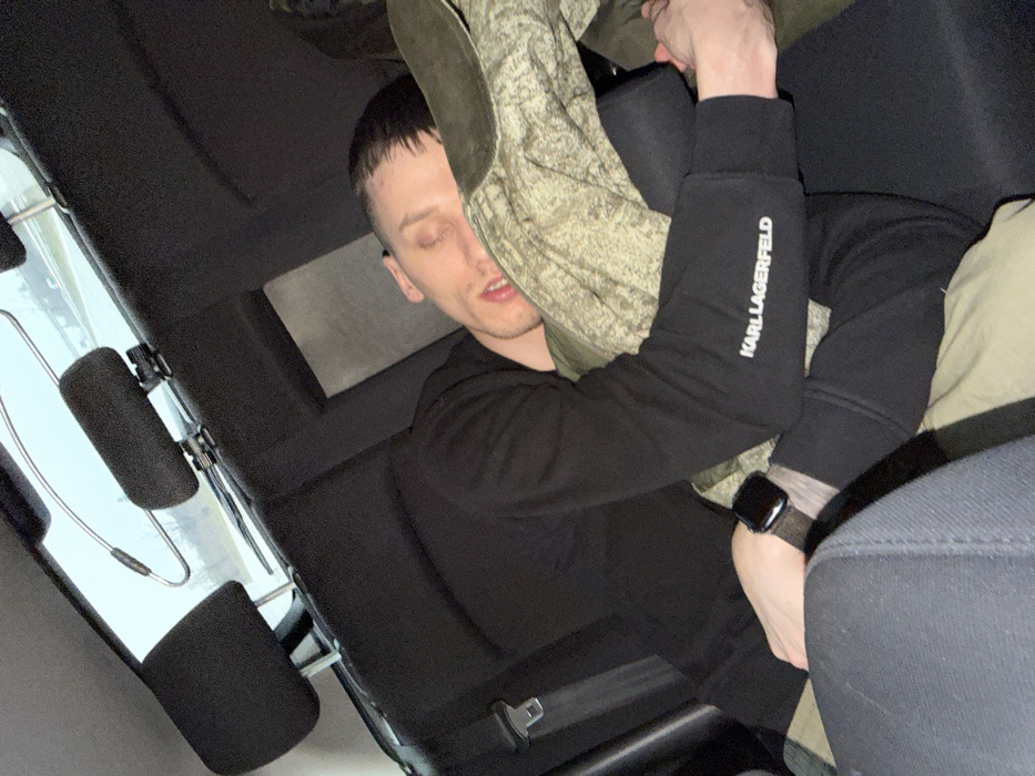 A male individual is depicted sleeping or resting in the rear passenger seat of a vehicle, in Gabarnitsa, Bulgaria. The subject, an adult male with short dark hair, has his eyes closed and mouth slightly ajar. He is wearing a black long-sleeved sweatshirt or hoodie with "KARL LAGERFELD" in white text visible on the left sleeve, and dark-colored trousers. A black smartwatch with a dark-textured band is present on his left wrist. His head rests on an olive green or camouflage-patterned fabric item, which also partially covers his lower body. The vehicle's interior features black fabric upholstery on the seats and headrests. Through the rear window, an indistinct outdoor scene is visible, suggesting a road or highway with guardrails and vegetation under an overcast sky. A silver-colored metal hanger-like object is suspended near the rear window.