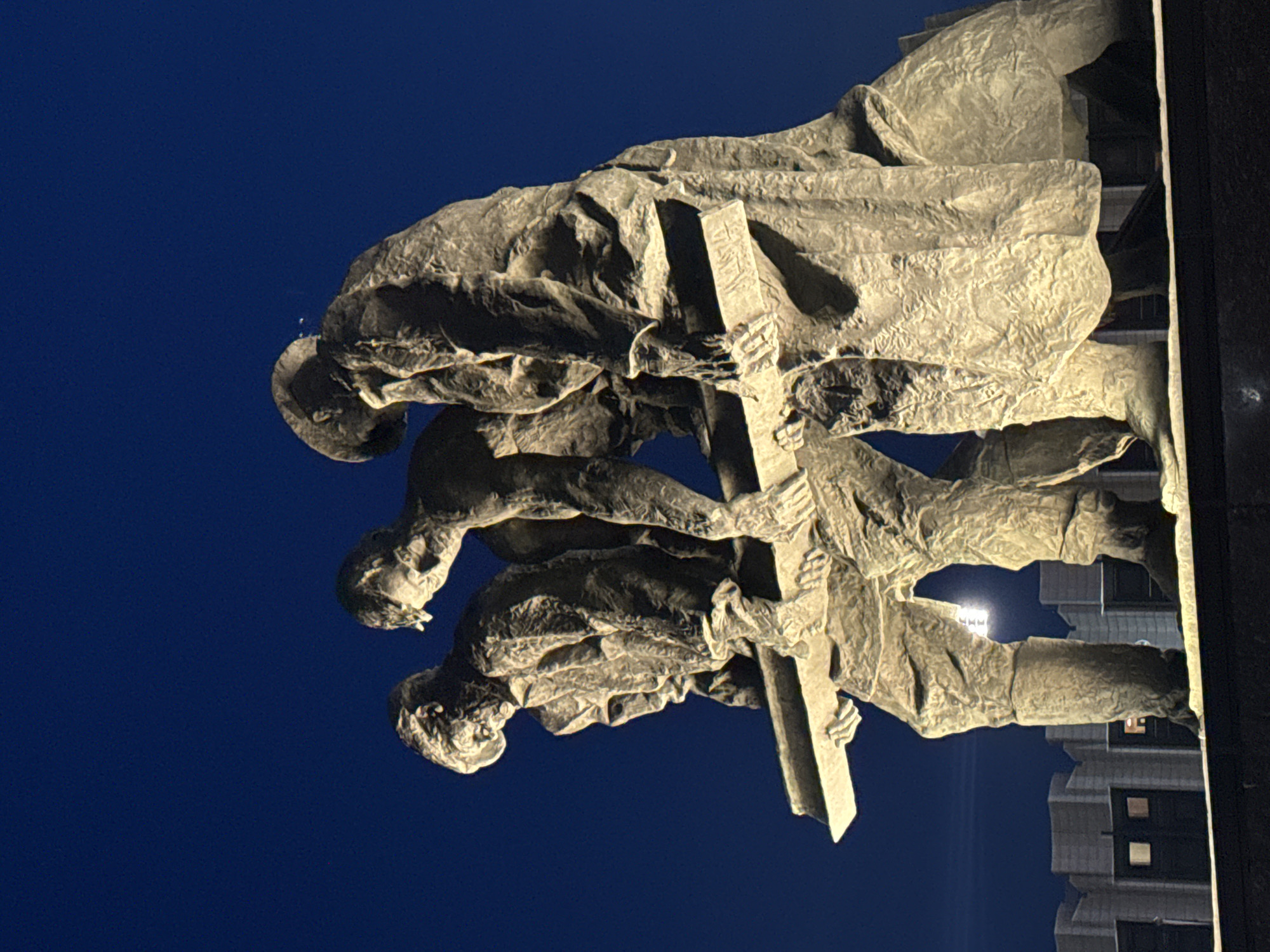 The image displays a large-scale bronze sculpture at night, depicting three male figures jointly carrying a substantial rectangular beam across their shoulders and hands. The figures are rendered with textured surfaces, suggesting heavy, worn clothing and determined expressions, with their gazes directed forward or downward. The monument is illuminated from below and the side, creating strong contrasts of light and shadow that emphasize its rugged details against a deep, uniform dark blue night sky. In the lower background, several dimly lit, block-like buildings with rectangular windows are visible, indicating an urban setting. A bright, square light fixture is discernible below and between the figures' legs. This monument, known as "To the Heroic Defenders of Leningrad," is located on Ploshchad Pobedy (Victory Square) in Saint Petersburg, Russia.