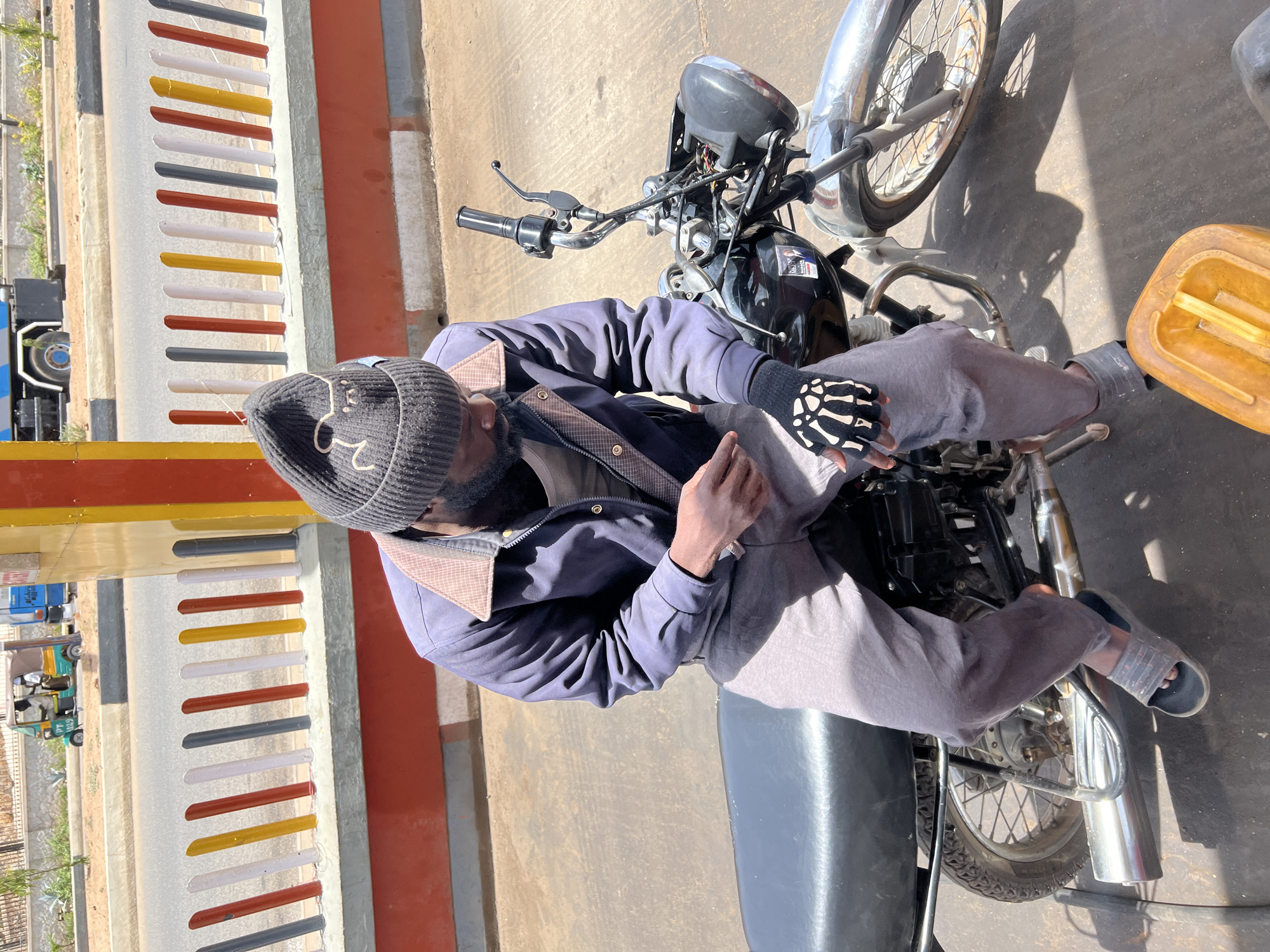 A man sits on a black motorcycle under bright, direct sunlight in what appears to be an outdoor, roadside setting in Jos, Nigeria. He is dressed in a dark blue jacket, light grey pants, a grey beanie hat with a white design, and clear plastic sandals. On his left hand, he wears a black fingerless glove featuring a distinctive white skeleton bone pattern.
The motorcycle is black with chrome elements, and a yellow plastic jerry can, possibly for fuel, rests on the ground beside it. Strong shadows are cast on the light-colored concrete surface, indicating mid-day sun.
Behind the man and motorcycle, a multi-colored barrier or fence stretches vertically, constructed from alternating horizontal bars of red, white, yellow, and dark grey. Beyond this fence, other vehicles are visible, including a blue and white bus and a green auto-rickshaw (keke Napep) with the text "JPT 029" on its side. The scene conveys a typical sunny day in an active, urban or semi-urban environment.