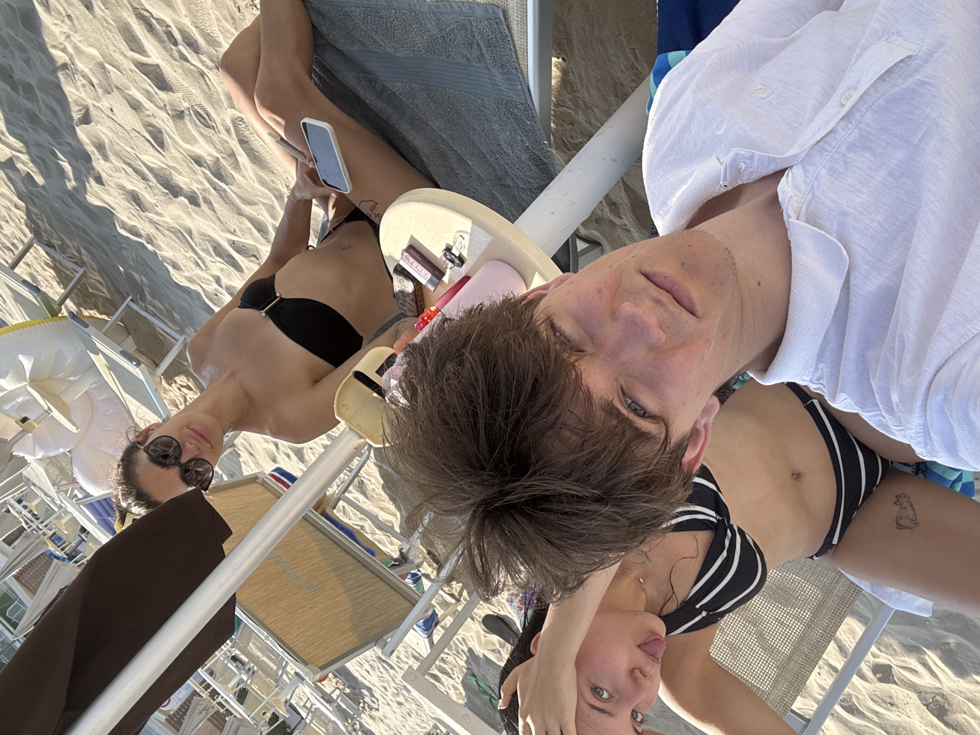 Three individuals are on a sandy beach, likely in Cupra Marittima, Italy, as suggested by the context. The person in the foreground is a young man with brown hair wearing a white polo shirt, holding a phone and taking a selfie. To his left, a young woman with dark hair is lying down, wearing a black and white striped bikini, and sticking out her tongue playfully. Further to the right, another young woman with dark hair tied up is wearing sunglasses and a black bikini top, also lying down. She is holding a smartphone. Beach chairs and umbrellas are visible in the background, indicating a typical beach resort setting. The overall atmosphere appears relaxed and casual.