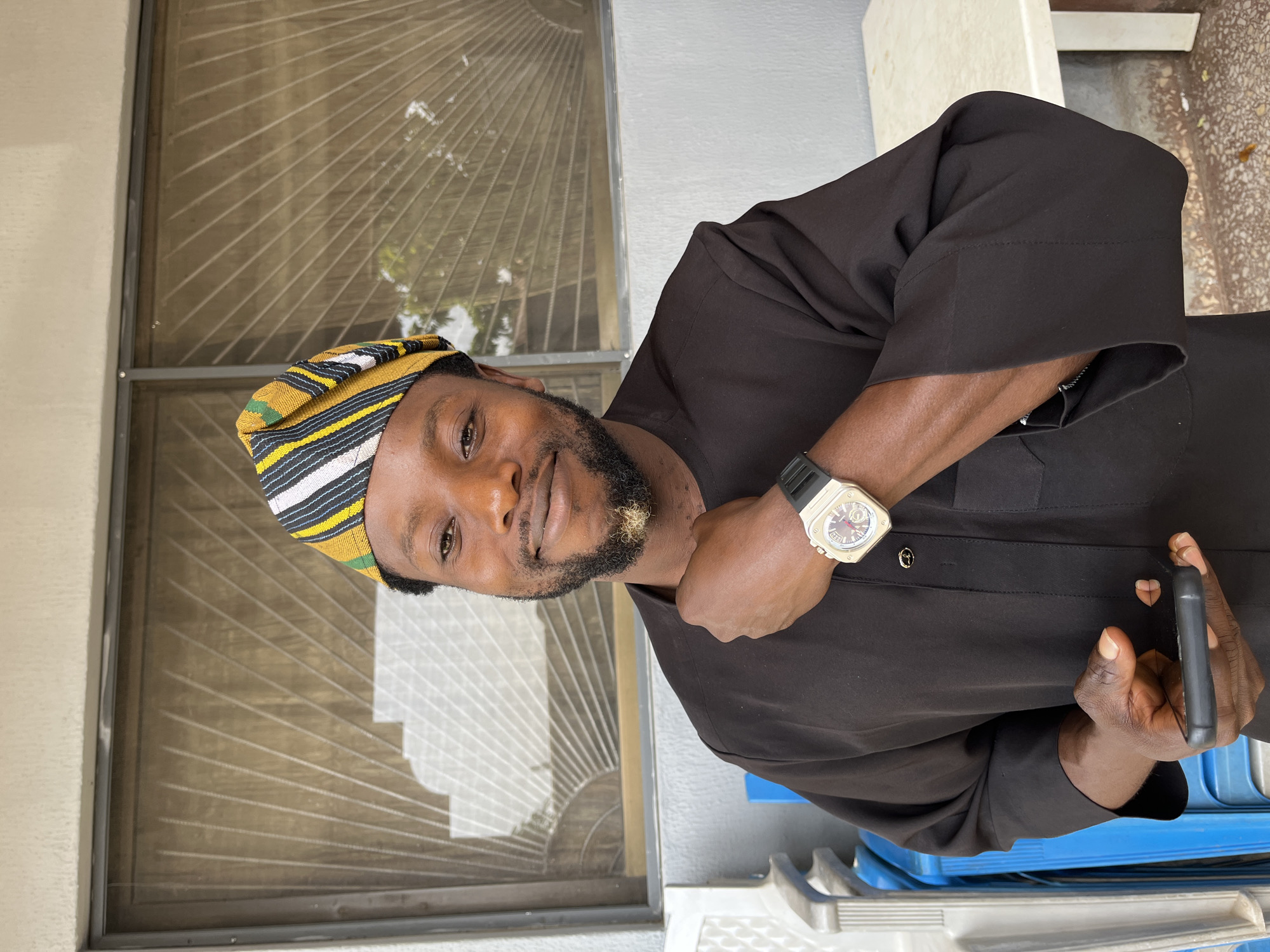 A medium shot captures a Black man standing outdoors, facing forward. He is wearing a dark brown, loose-fitting traditional outfit and a patterned cap in yellow, black, white, and green stripes. His left hand is resting on his chest, near his chin, and he is wearing a silver wristwatch with a black band on his left wrist. He holds a dark smartphone in his right hand, partially visible near his chest. The man has a beard, and he is smiling. The background features a window with blinds and a beige wall. To the left of the frame, blue plastic chairs and a metal railing are visible, and to the right, part of a white table is present. The ground beneath is paved with reddish-brown stones. The scene is likely an outdoor courtyard or patio area in Jos, Nigeria.
