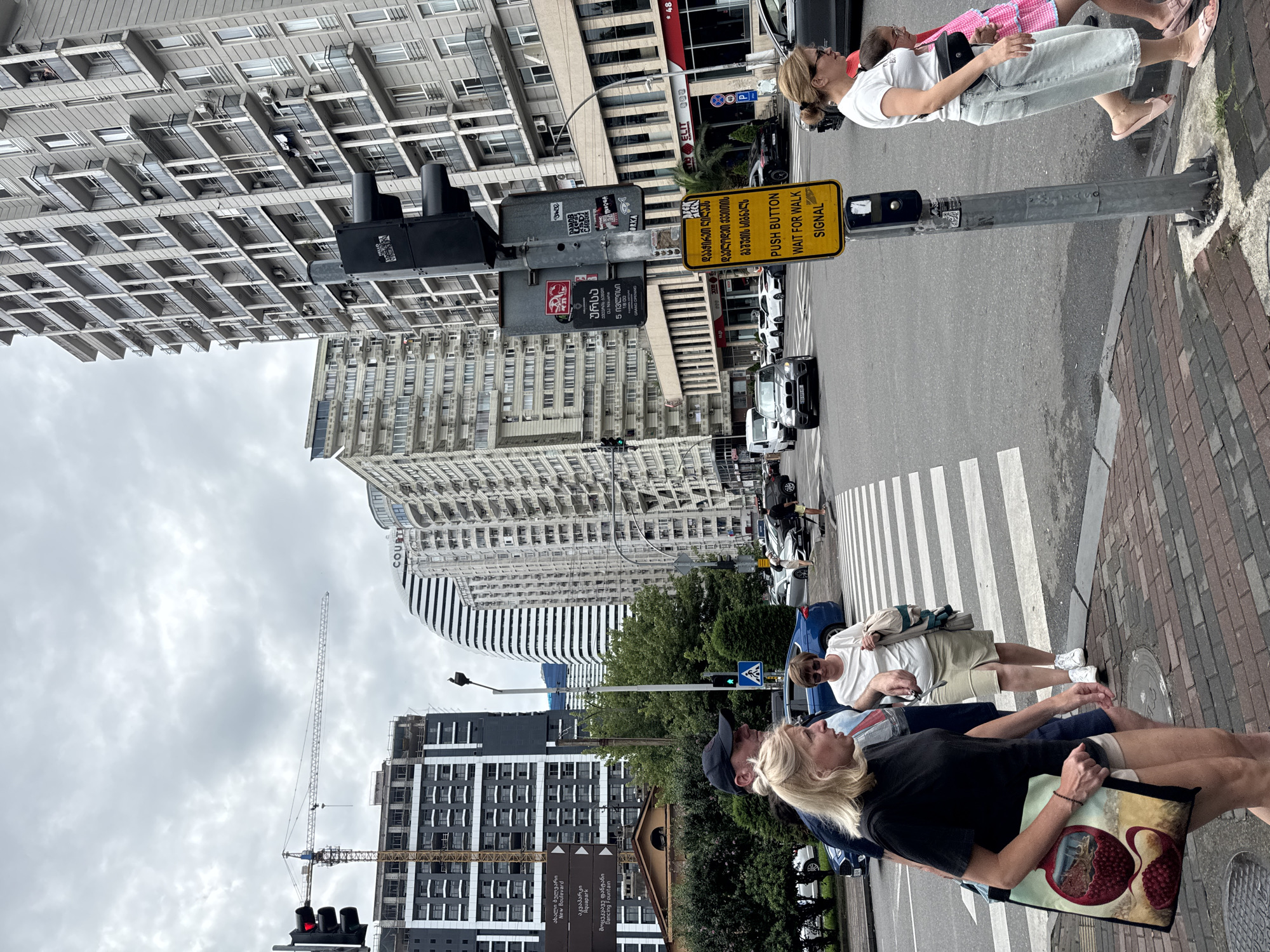 The image shows a street scene in Batumi, Georgia, on a cloudy day. In the foreground, several people are crossing a crosswalk. On the left, a woman with blonde hair in a black t-shirt and beige shorts is carrying a large tote bag with a pomegranate design. Beside her, a man wearing a dark baseball cap and a blue t-shirt is walking. Behind them, a woman in a white t-shirt and beige skirt is walking across the street. To the right, a woman in a white t-shirt and light blue jeans is walking with a young girl in a pink dress.
The background features tall buildings, including a modern, curved white building with a striped facade, and other multi-story residential buildings. A construction crane is visible in the distance, indicating ongoing development. Traffic lights and street signs are present, including a yellow sign that reads "PUSH BUTTON WAIT FOR WALK SIGNAL" in English and Georgian. Cars are visible on the street, including a black SUV and a blue sedan. The street is paved with cobblestones in the foreground and asphalt further back.
