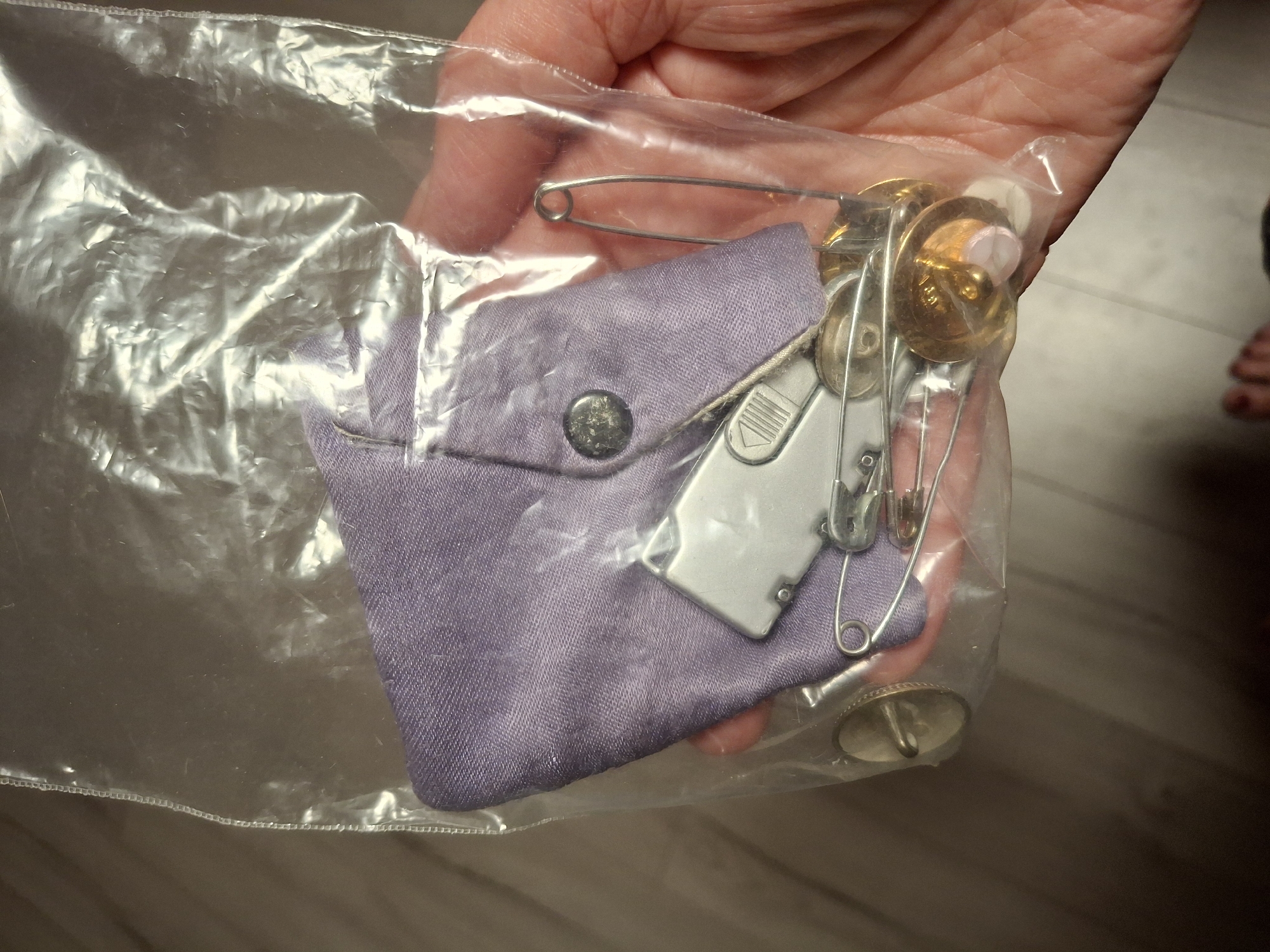 The image shows a close-up view of a small sewing kit in a clear plastic bag. The kit contains a folded piece of lavender-colored fabric, a small silver folding utility knife, several safety pins of various sizes, two round brass buttons with what appears to be pink thread or fabric underneath, and a single brass button with a silver pin attached. The kit is being held by a person's hand, which is visible on the right side of the frame. The background is out of focus and appears to be a wooden floor. The image is taken from a slightly elevated angle, looking down into the bag. There is no discernible information in the image that places the scene in Ogre, Latvia.