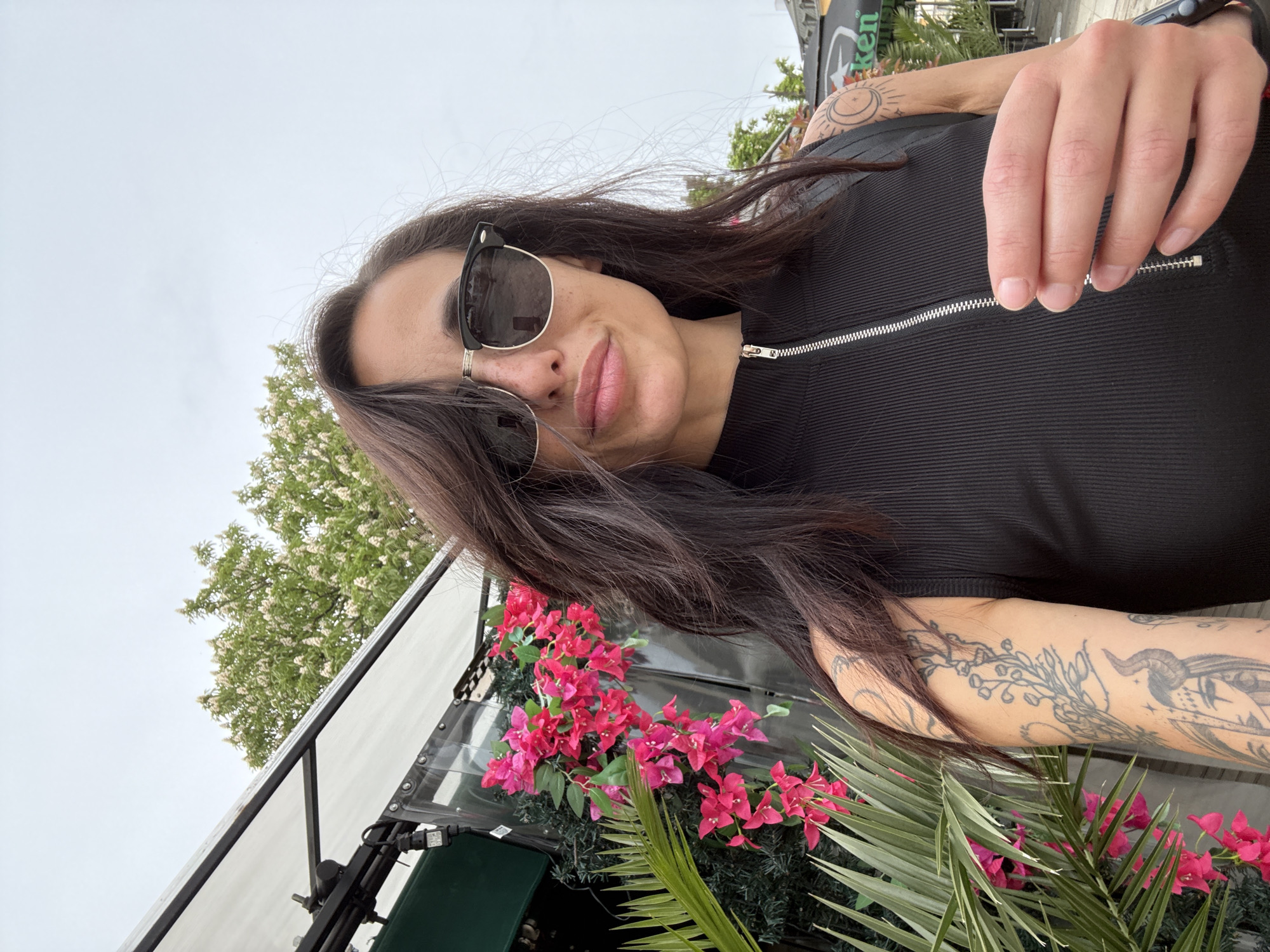 A woman with sunglasses and long brown hair is pictured from a low angle. She is wearing a black, short-sleeved top with a zipper. Her right arm is visible, featuring multiple tattoos. In the background, there are pink flowers and palm leaves, along with a white awning structure. A tree is visible behind the awning. The city of Mezdra, Bulgaria, cannot be confirmed from the provided image.