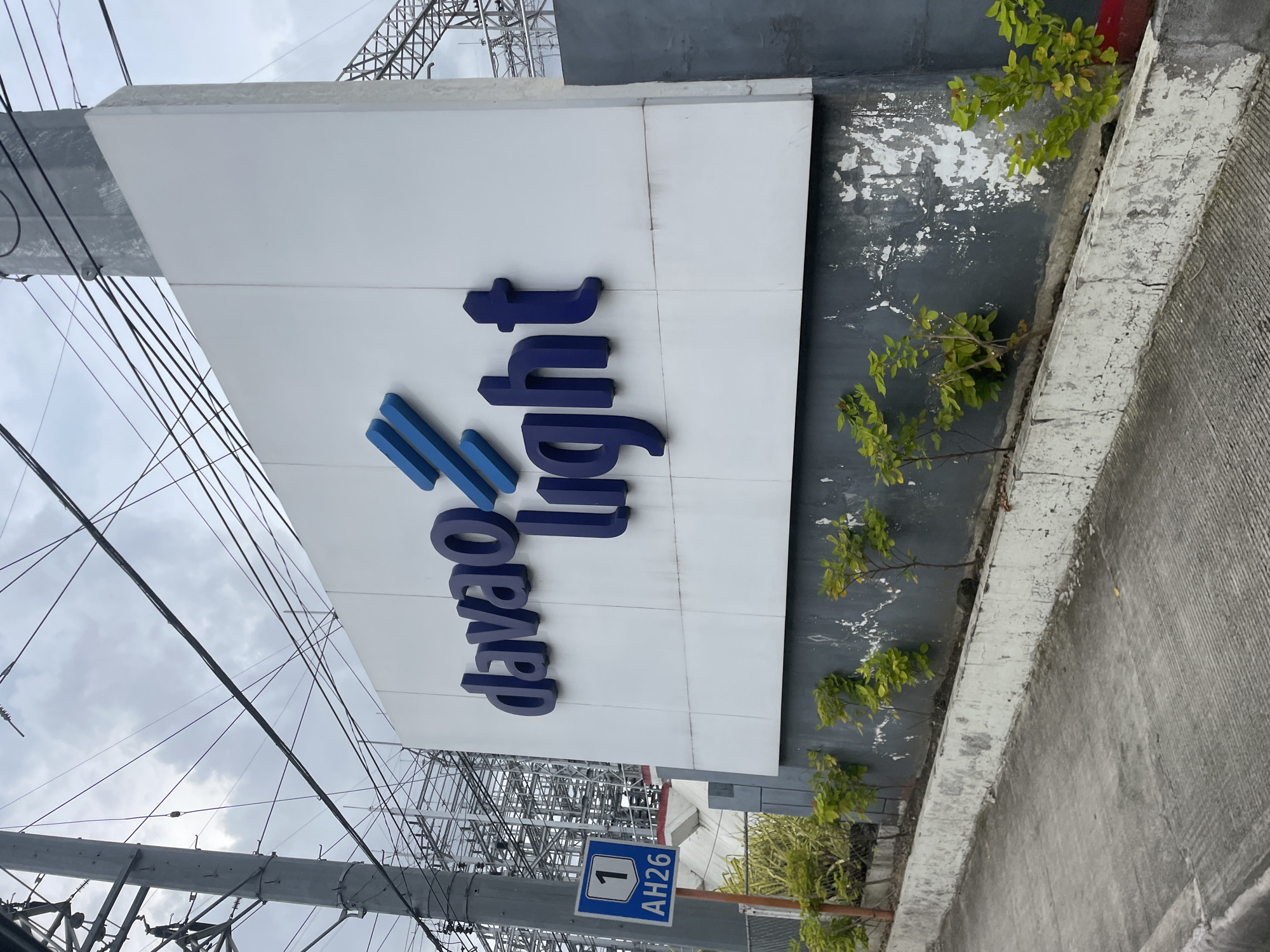 The image depicts the sign of "Davao Light" which is a power distribution utility company serving Davao City, Philippines. The sign is white with blue lettering and blue geometric shapes. In front of the sign, there is a concrete barrier with small trees and bushes planted along it. To the left of the sign, a blue and white road sign with the number "1" and "AH26" is visible. The background shows utility poles and electrical infrastructure, suggesting the location is near a power substation or facility. The sky is overcast. The overall scene is an exterior view of a corporate sign in an urban environment.