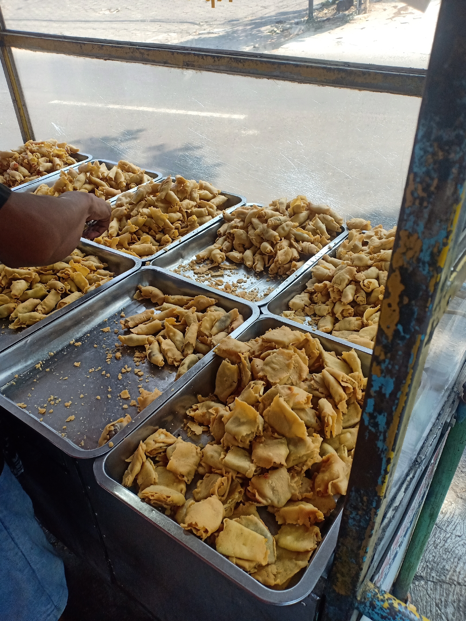 This image depicts a street food stall in Semarang, Indonesia, showcasing a variety of fried snacks. The stall is set up with multiple metal trays filled with golden-brown, deep-fried items. In the foreground, a person's hand, with dark skin and wearing a dark sleeve, is reaching into one of the trays, likely selecting or serving food. The trays are arranged in a grid-like fashion, with some containing smaller, cylindrical fried pieces, and others filled with larger, flatter pieces. Crumbs and remnants of the fried food are scattered across the metal surfaces of the trays and the stall. The stall itself appears to be a mobile cart with a metal frame, some parts of which are painted with yellow, blue, and green, showing signs of wear and tear. Behind the food, a transparent or semi-transparent barrier, possibly plastic sheeting, separates the food from the exterior, revealing a blurred background of a street with visible road markings and some distant structures, indicating an urban setting. The lighting suggests it is daytime, possibly morning or afternoon, with sunlight illuminating the scene.