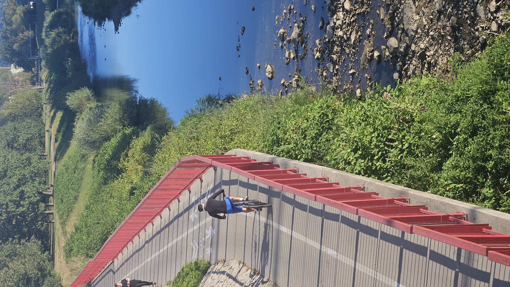 This image shows a scenic view of the Sekčov river in Prešov, Slovakia, as seen from a pedestrian and bicycle bridge. The bridge, with its distinctive red railing, runs along the left side of the frame, curving gently. On the bridge, a person is cycling away from the camera, wearing a dark long-sleeved shirt, blue shorts, and a helmet. Further up the bridge, another person in dark clothing is walking, also away from the camera. The bridge path is marked with white lines, including cycle path symbols.
The Sekčov river flows on the right side of the image. The water is a deep blue, reflecting the clear sky. The riverbanks are lined with lush green vegetation, including trees and bushes, creating a vibrant natural setting. The right bank of the river features a rocky shoreline with numerous stones of various sizes scattered in the shallow water and along the edge. The overall scene suggests a peaceful day, ideal for outdoor activities like cycling and walking, with the river and its surroundings bathed in bright sunlight.